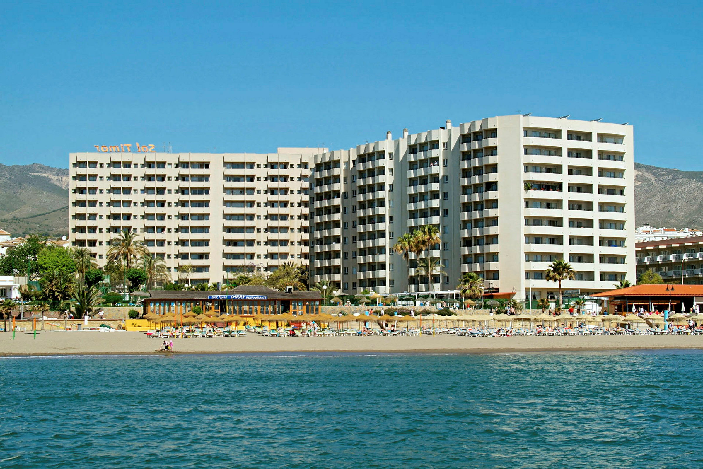 Strandhotell med høye bygninger og solsenger ved vannet. (AI-generert tekst)