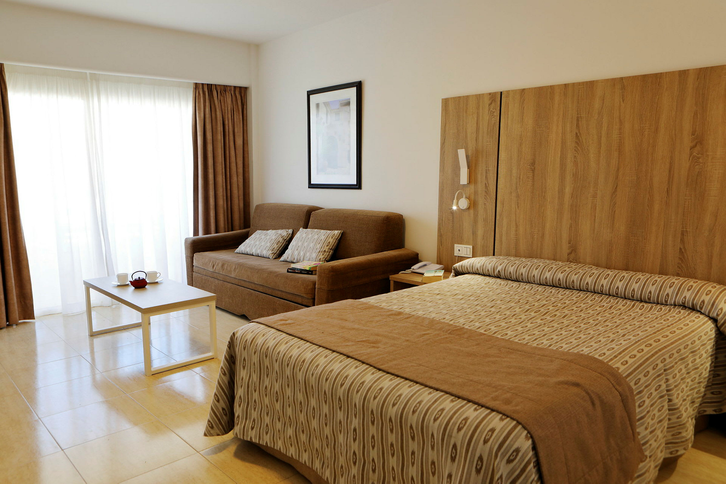 Hotellrom med en brun sofa, hvite gardiner, et bord og en seng. (AI-generert tekst)
