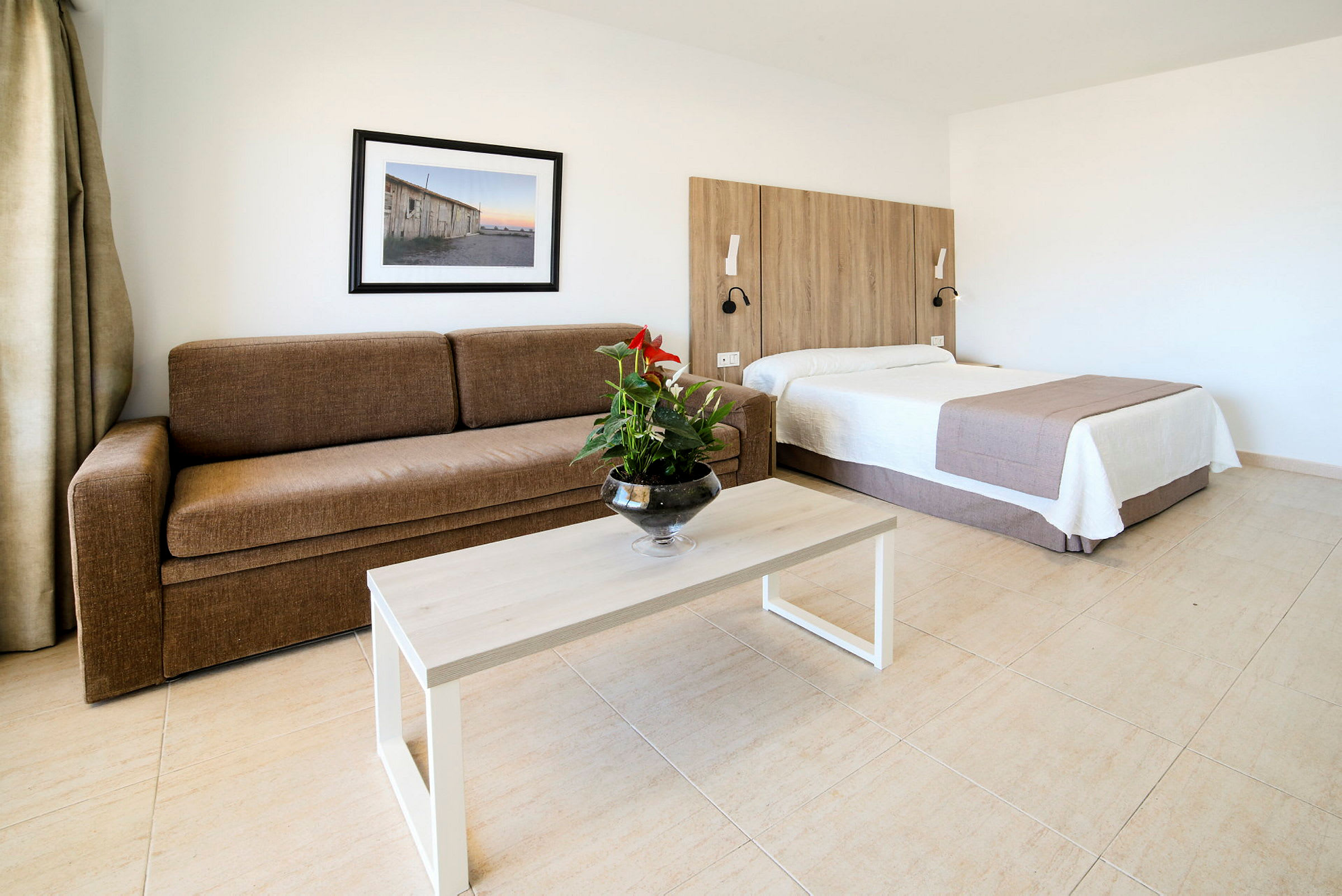 Et hotellrom med en brun sofa, et bord med en blomsterpotte, og en seng med trepanel bak. (AI-generert tekst)
