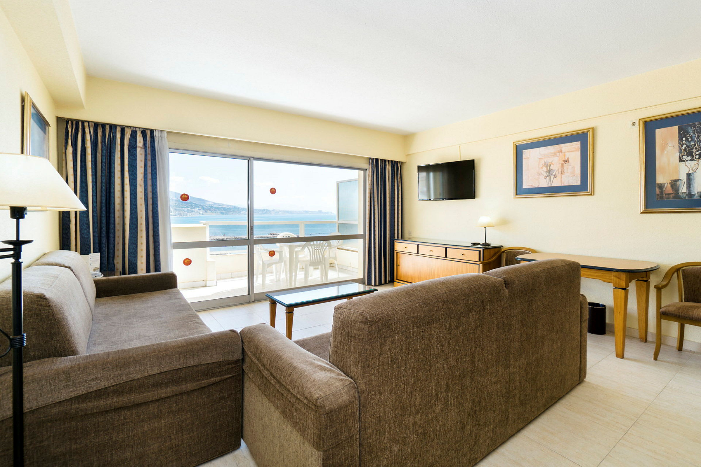 Stue med sofaer, bord, TV og utgang til balkong med utsikt over havet. (AI-generert tekst)