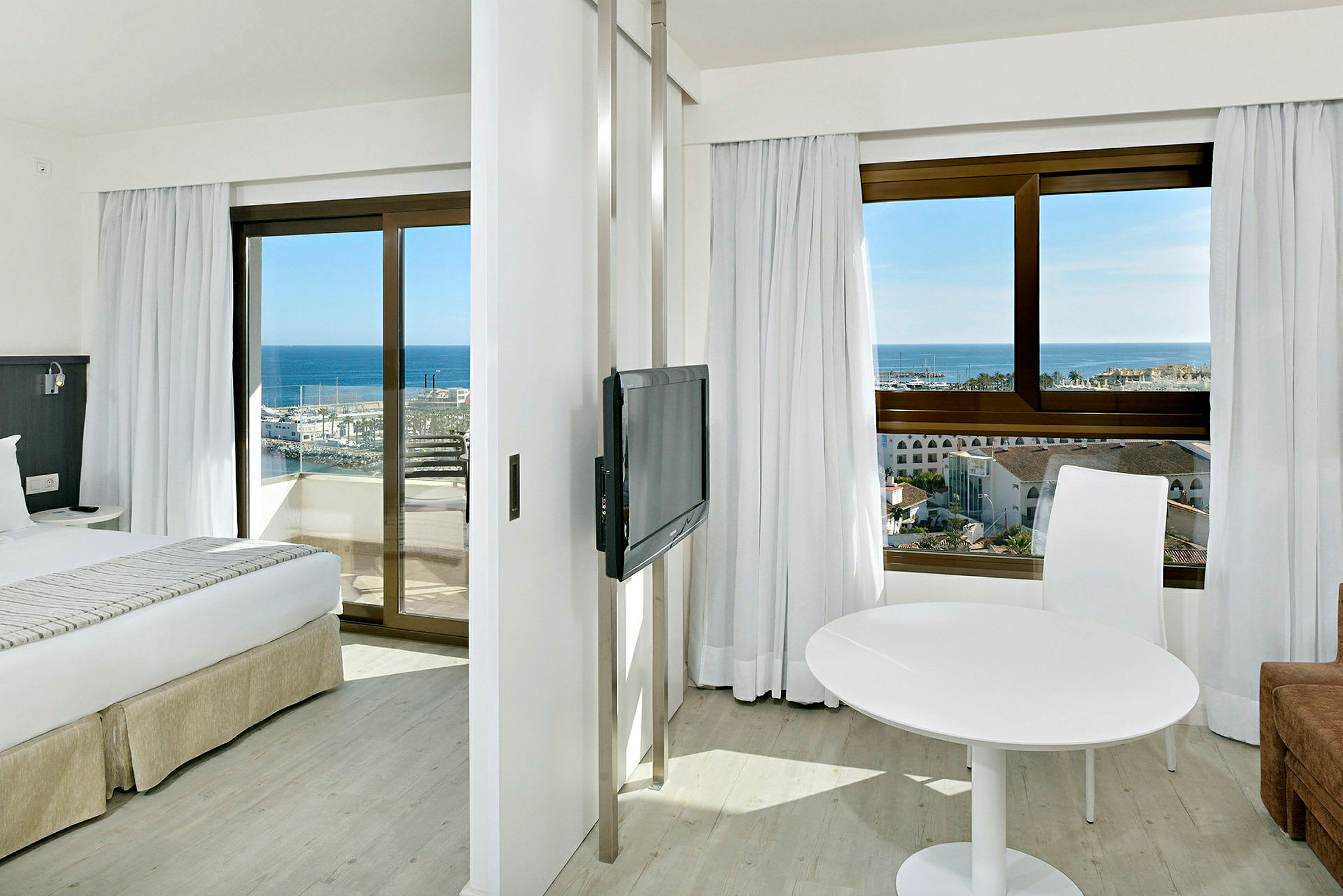 Hotellrom med to vinduer som vender mot havet, en seng, en TV og et hvitt bord med en stol. (AI-generert tekst)