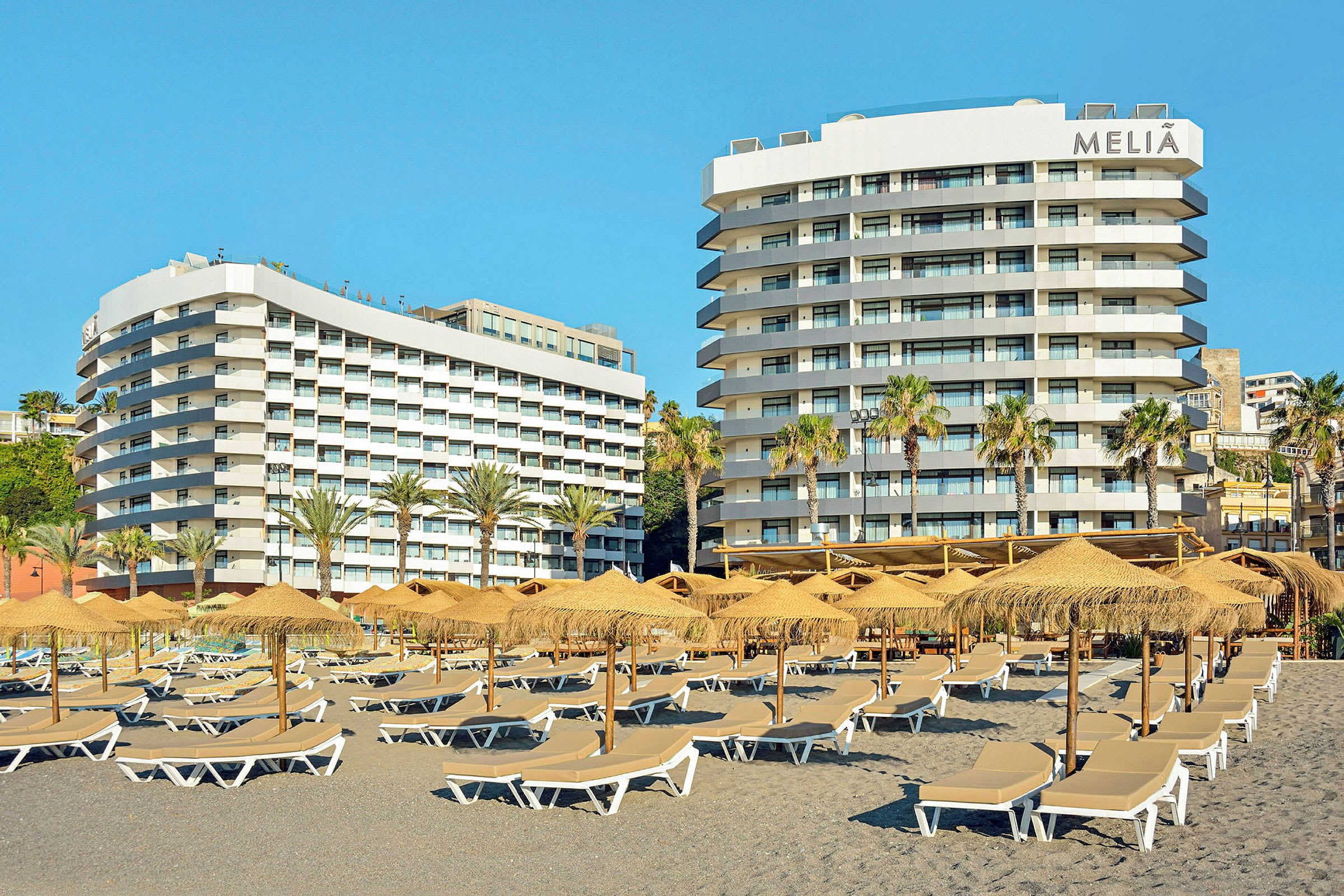 Bilde av to hotellbygninger med balkonger. Foran finnes parasoller og solsenger på en strand. (AI-generert tekst)