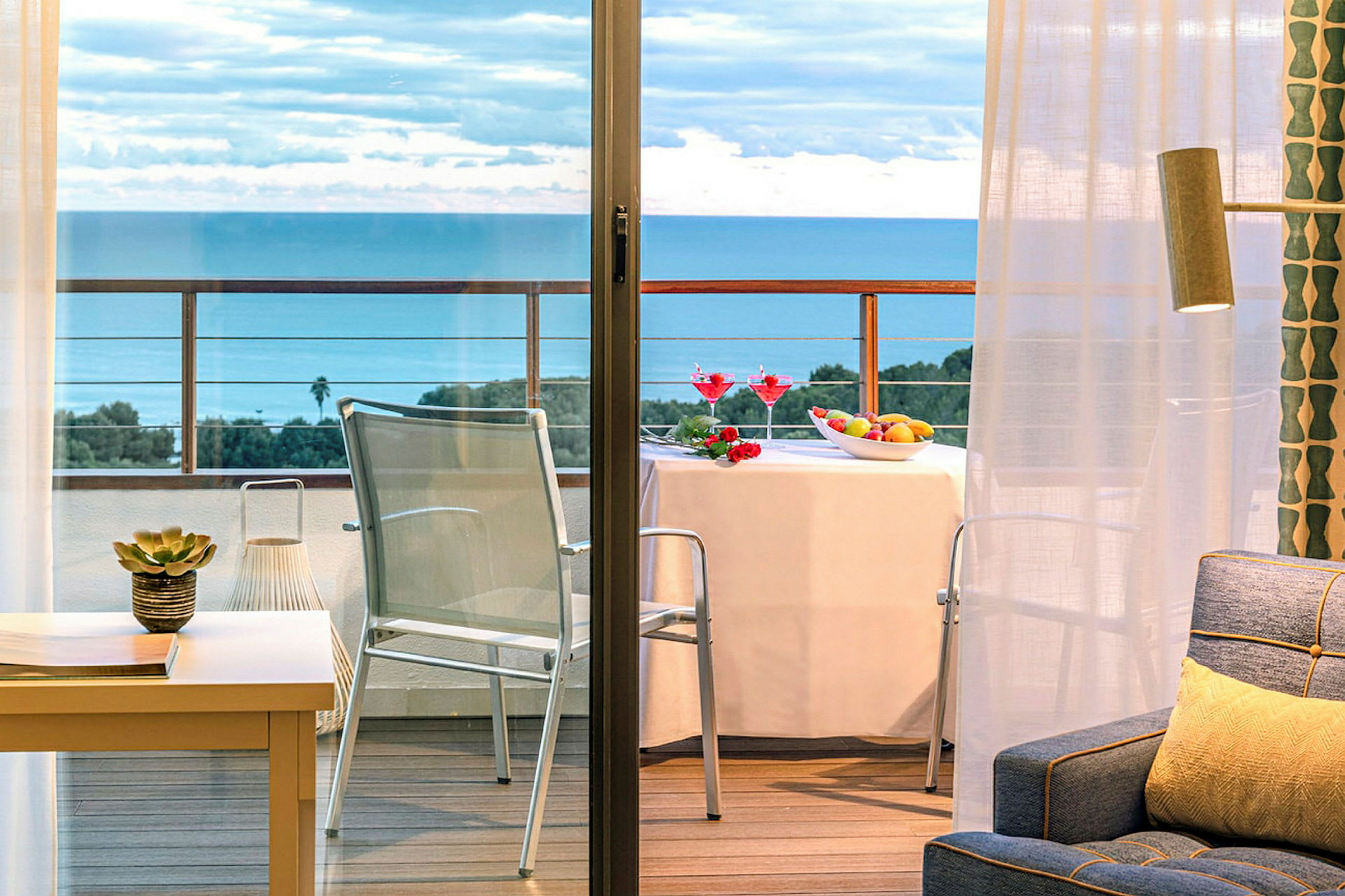En balkong med utsikt over havet, et bord med duk, frukt og drinker, og stoler. Innendørs er en sofa og dekorasjoner synlige. (AI-generert tekst)