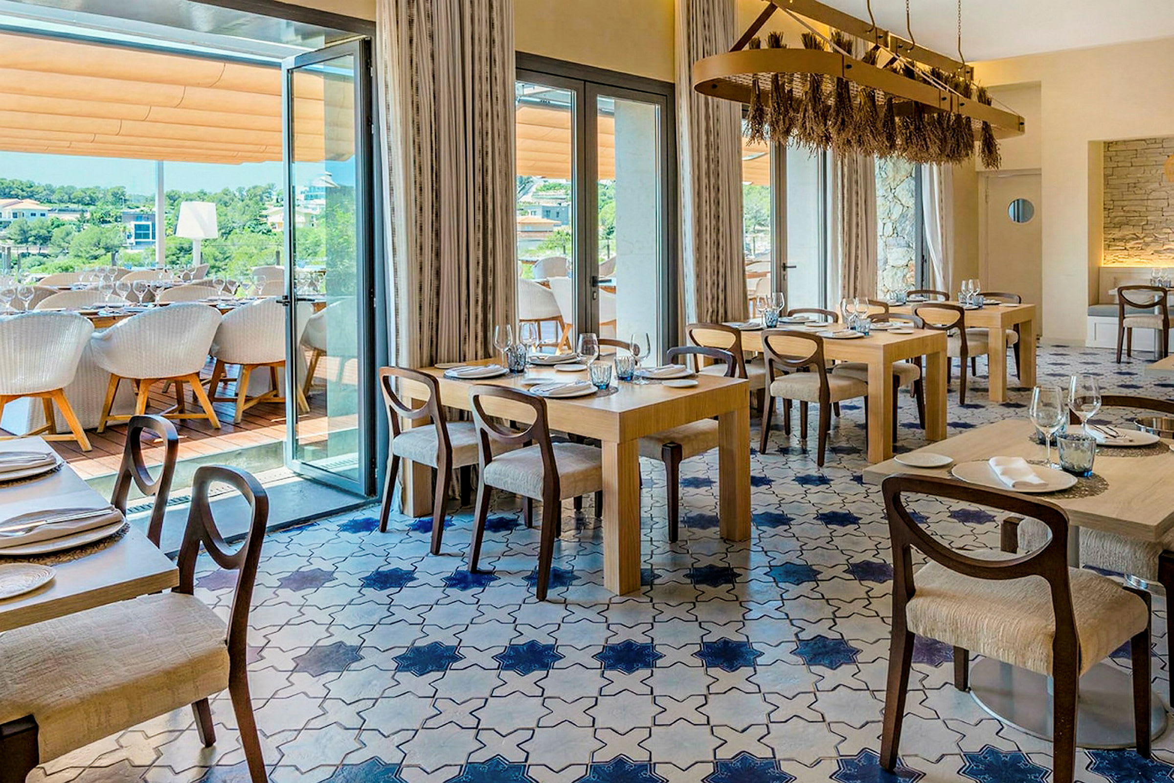 En elegant restaurant med flere bord og stoler, blått og hvitt flislagt gulv og store vinduer som fører ut til en terrasse. (AI-generert tekst)