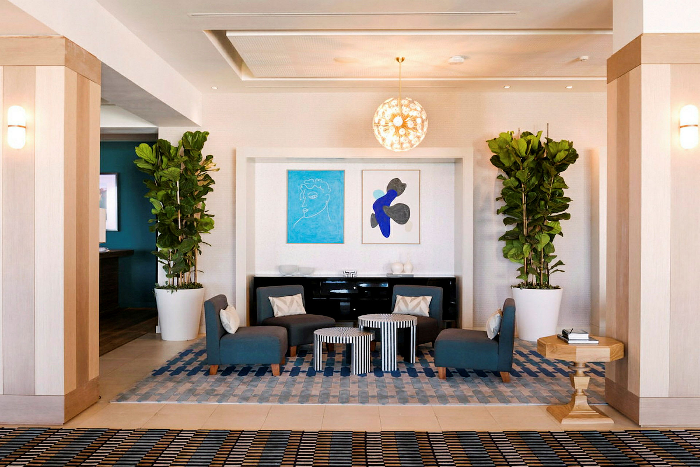 En hotellobby med sitteplasser i form av blå stoler rundt små bord. Planter i store hvite potter og kunstverk på veggen pynter rommet. (AI-generert tekst)