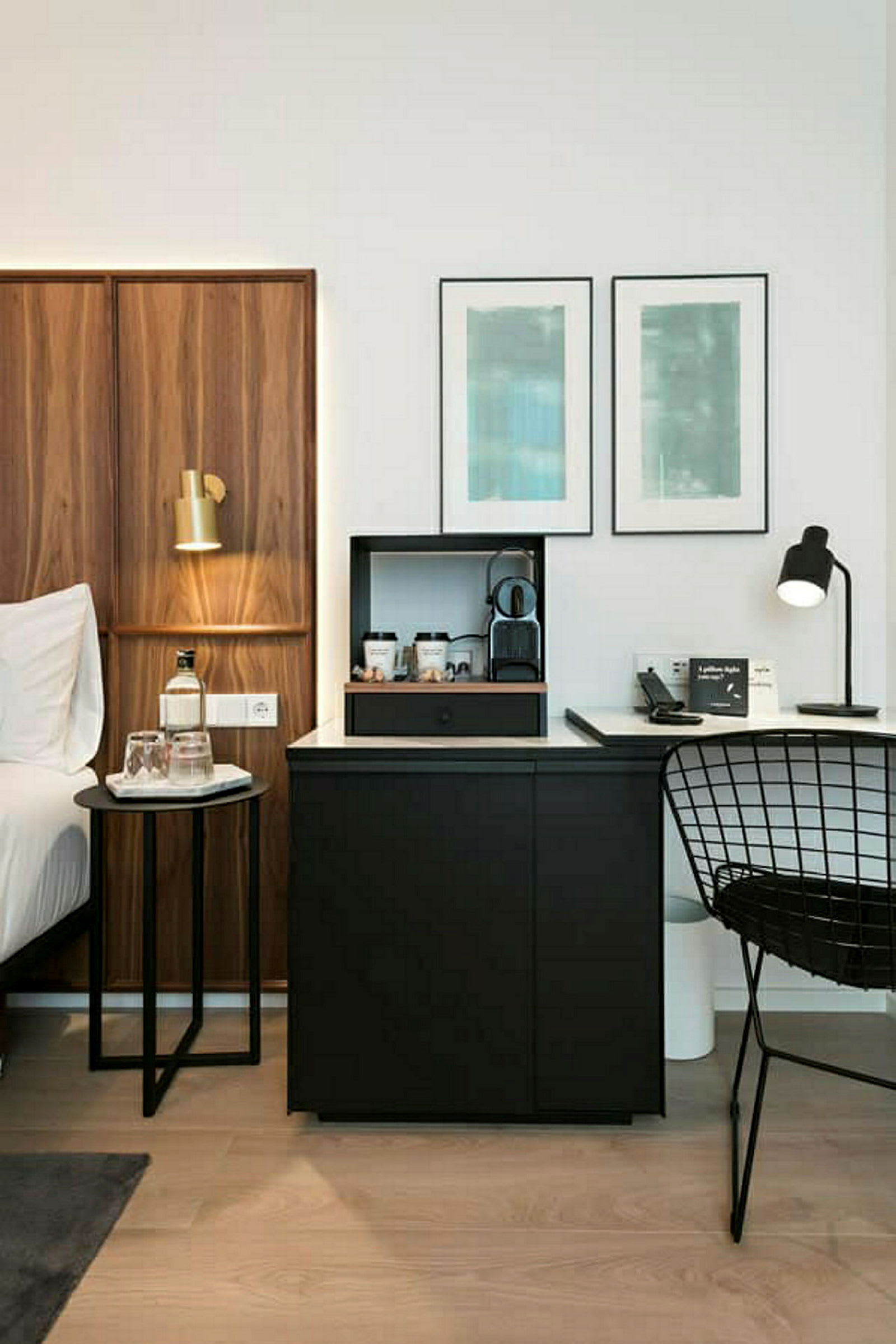 Et hotellrom med en seng til venstre, et lite bord med en flaske vann og glass. På høyre side er det et svart skrivebord med en kaffemaskin og en stol. (AI-generert tekst)