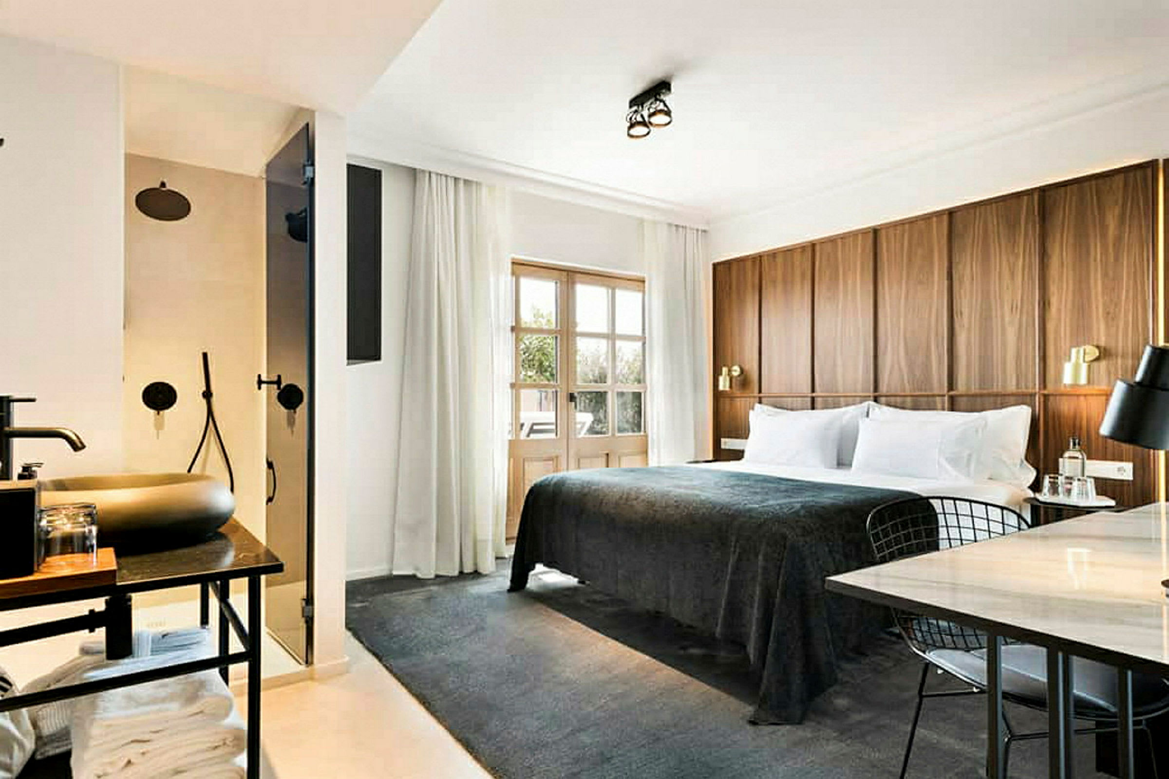 Et hotellrom med en stor seng med mørkgrå sengeteppe, en trepanel som sengegavl, et arbeidsområde med bord og stol, samt et synlig bad med dusj bak en glassdør. (AI-generert tekst)