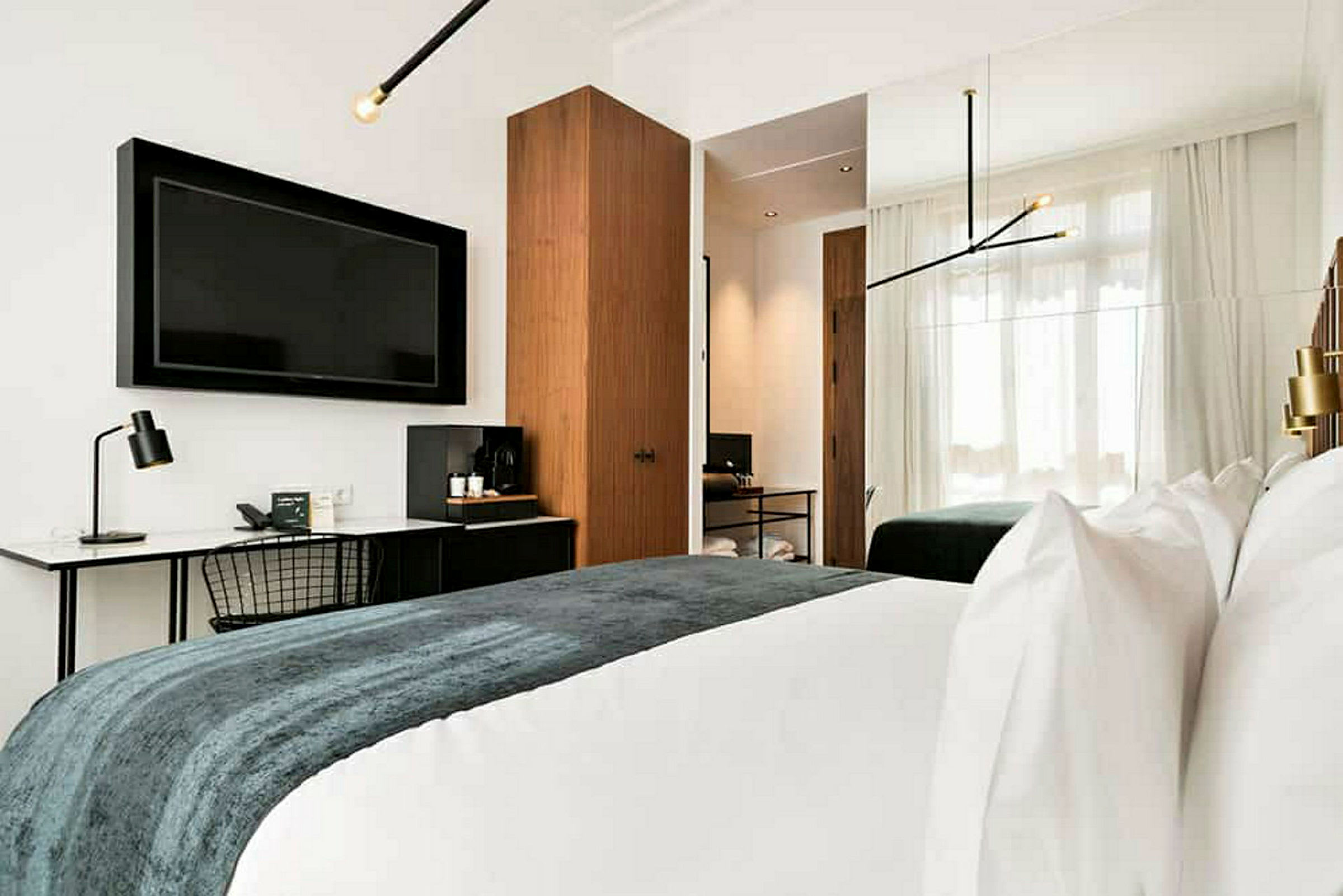 Bilde av et moderne hotellrom med en stor TV på veggen, skrivebord med lampe, en dobbeltseng med hvitt sengetøy og et teppe i mørke farger. Rommet har også et skap og store vinduer med lyse gardiner. (AI-generert tekst)