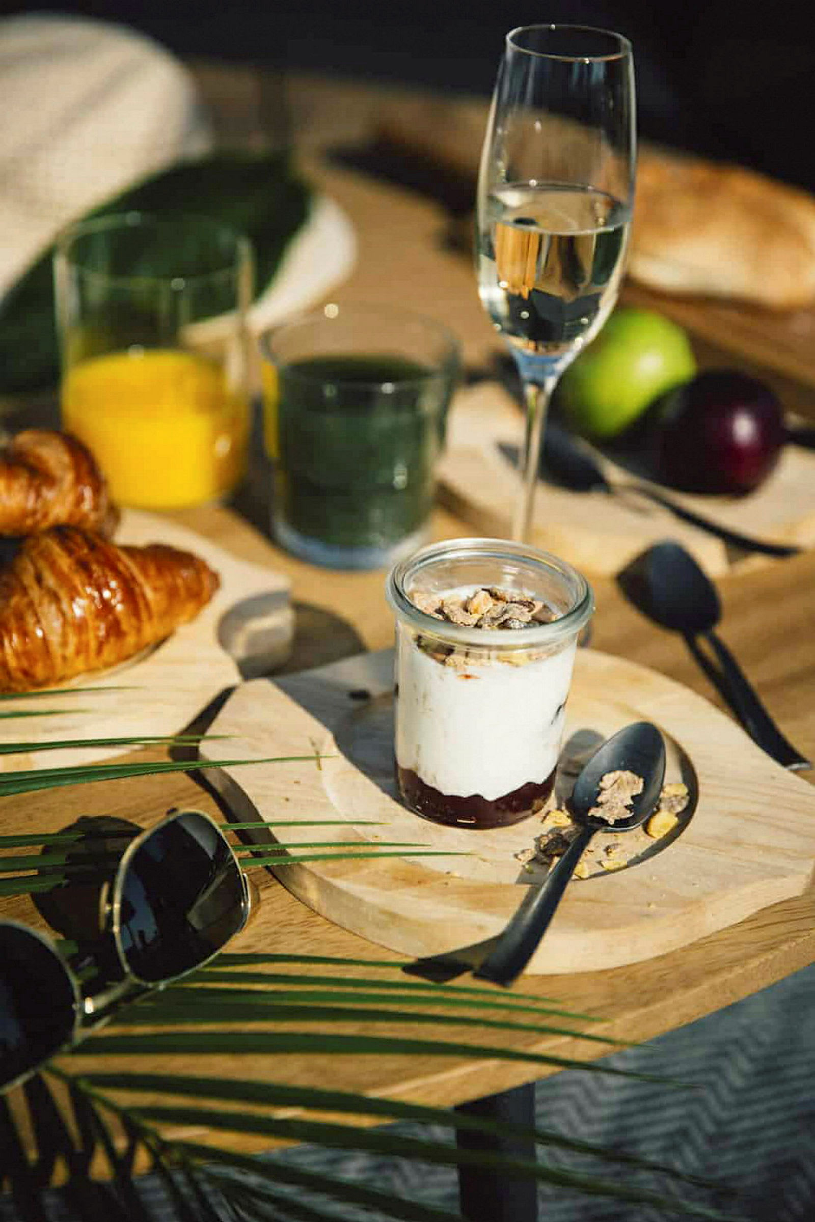 Et frokostbord med glass fylt med drikkevarer, en croissant, en tallerken med yoghurt og granola samt andre detaljer som briller og frukt. (AI-generert tekst)