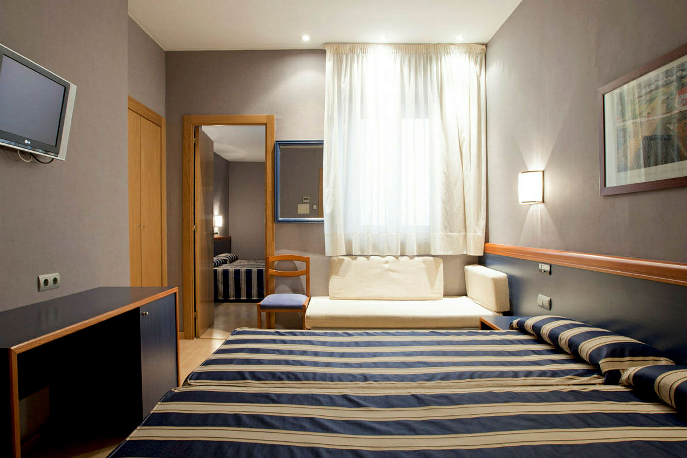 Et hotellrom med en oppredd enkeltseng med stripete sengeteppe, et skrivebord og en sitteplass ved et vindu med lyse gardiner. (AI-generert tekst)