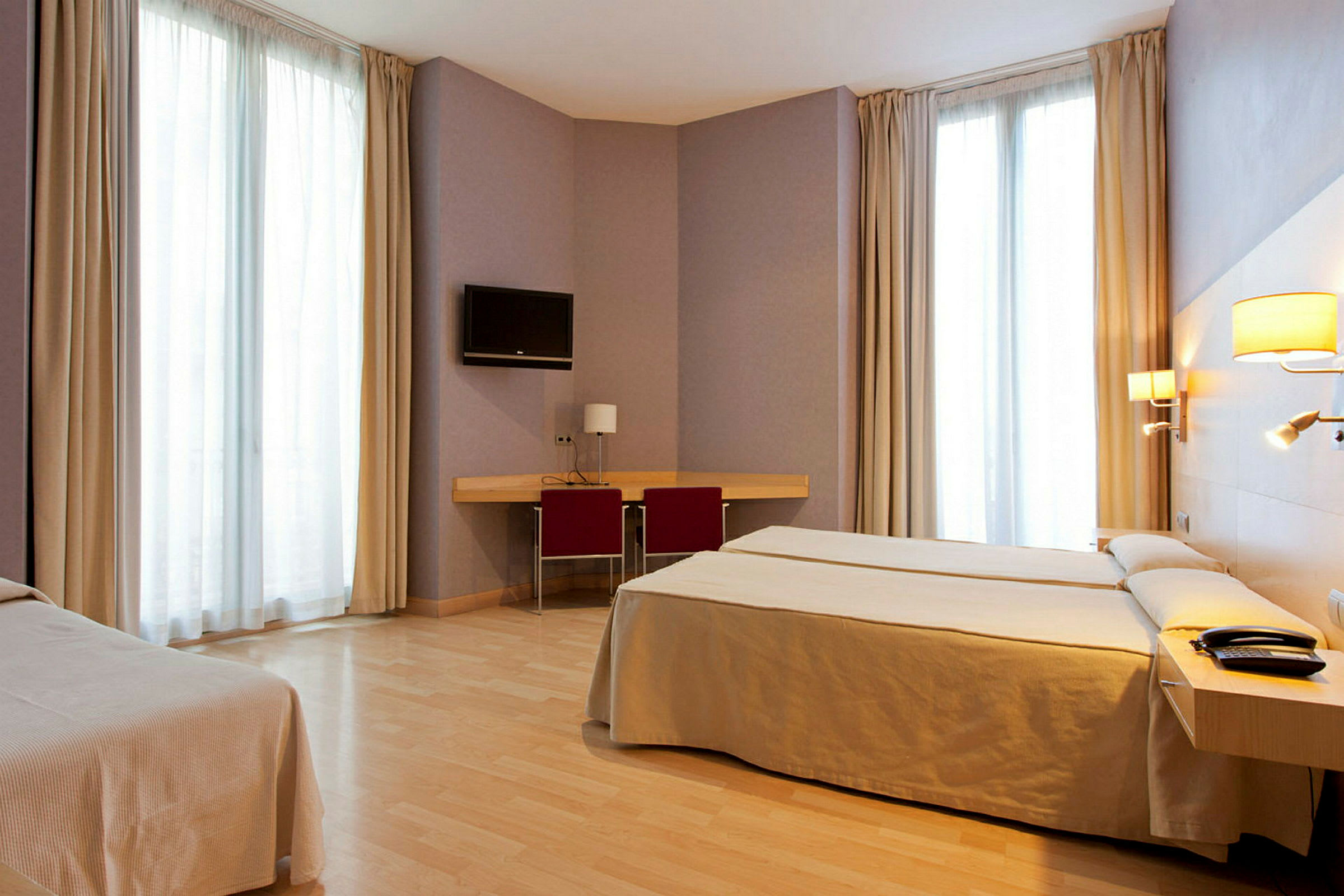 Et hotellrom med to senger, skrivebord med stol, veggmontert TV og store vinduer med gardiner. (AI-generert tekst)