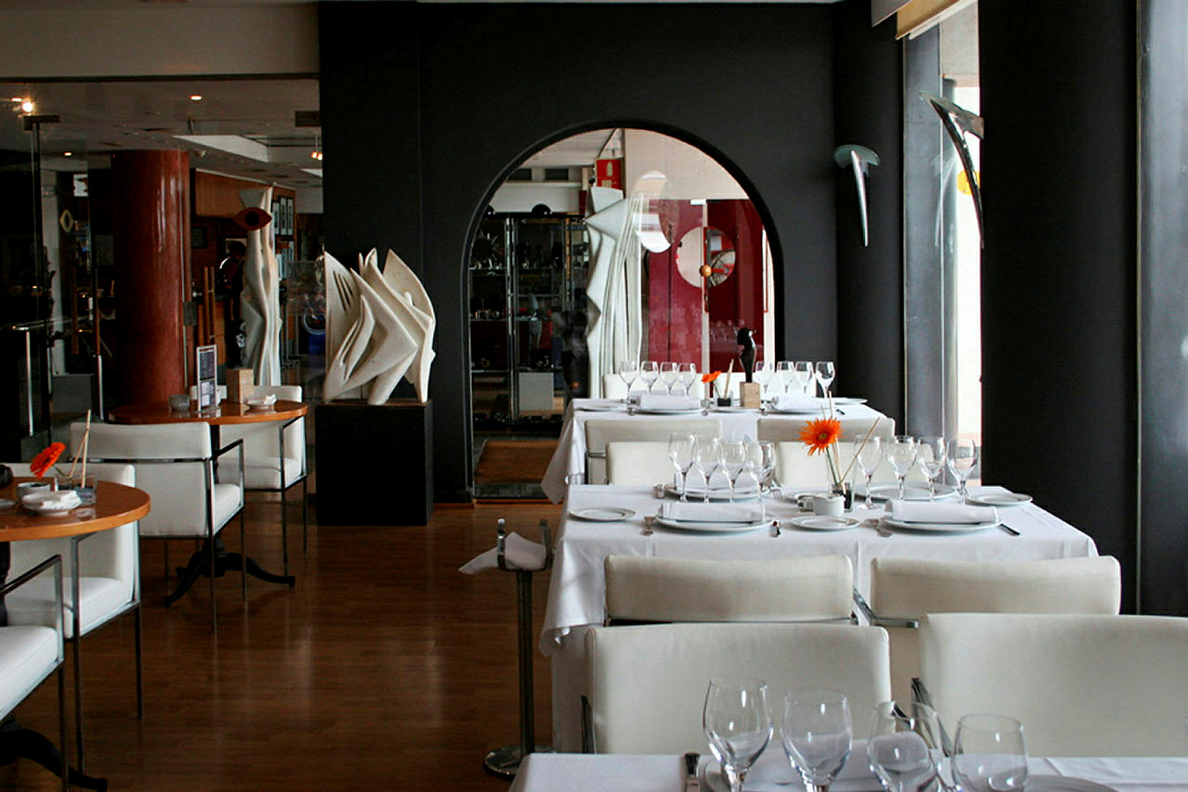 En restaurant med moderne designmøbler. Bordene er dekket med tallerkener, glass og bestikk. Veggene har kunstverk og dekorasjoner. (AI-generert tekst)