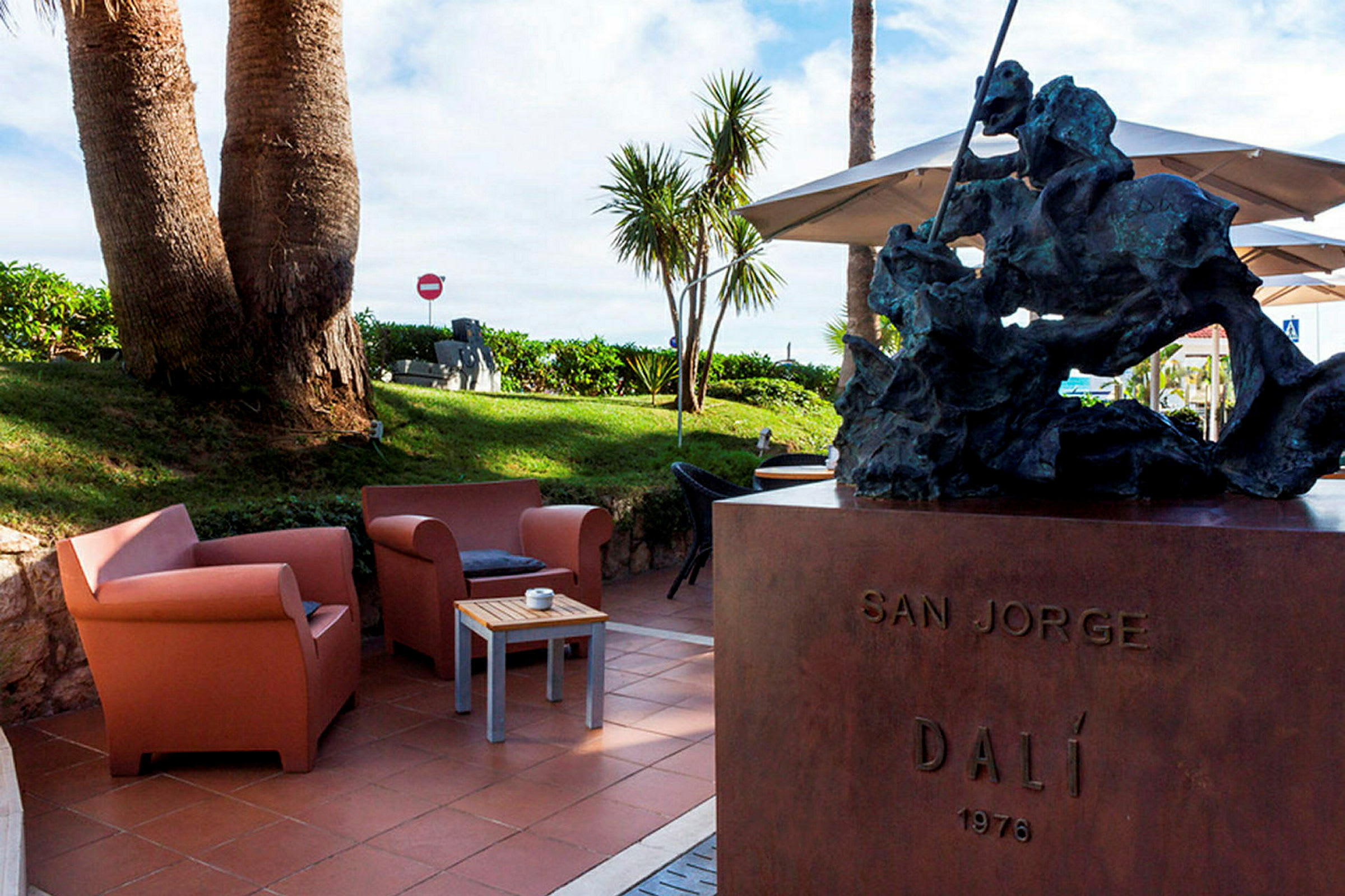 En statue står på en sokkel med teksten 'SAN JORGE DALÍ 1976'. I bakgrunnen sees en plen med palmer og sittemøbler på en terrasse. (AI-generert tekst)