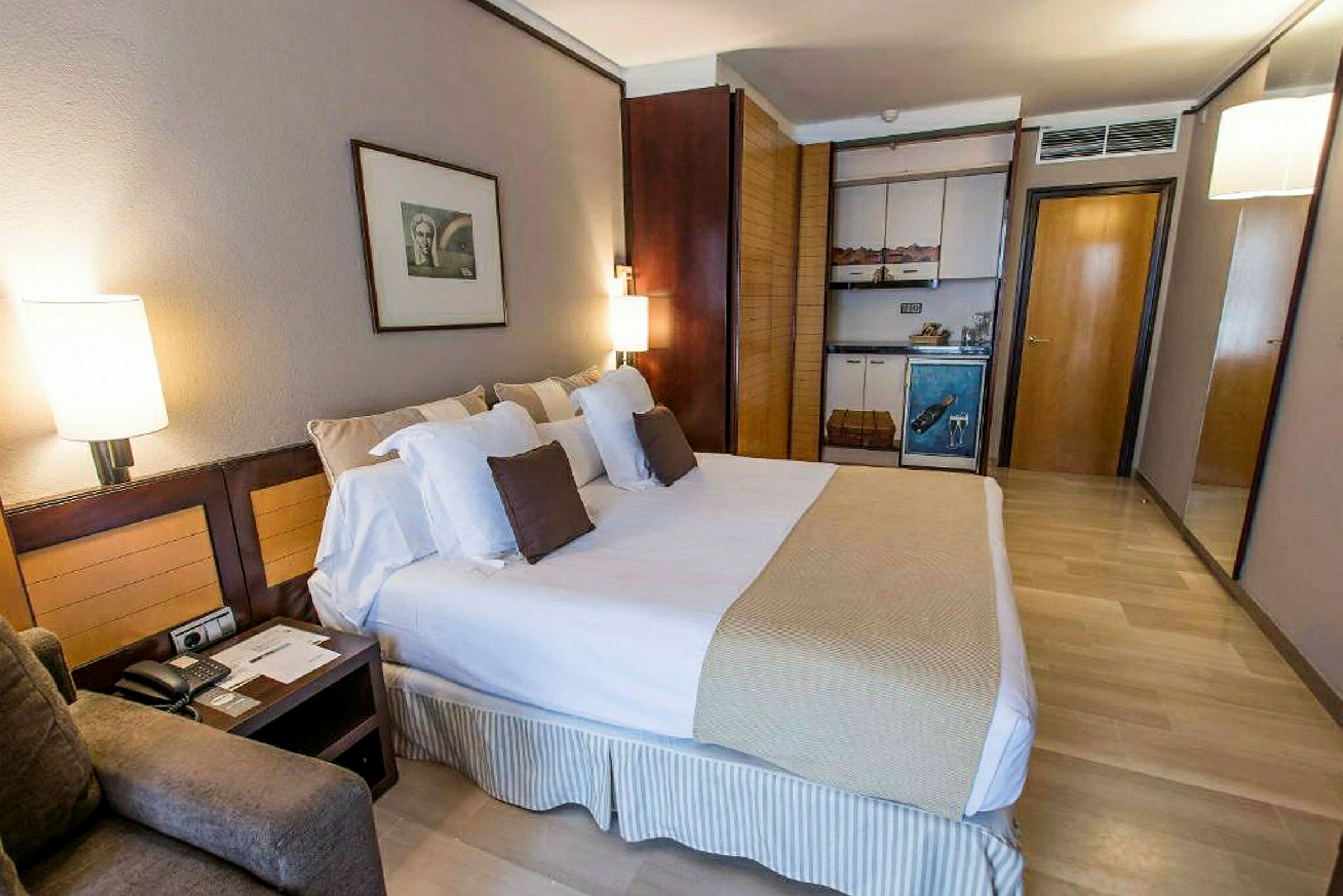 Et hotellrom med en stor seng, puter, nattbord og en liten kjøkkenkrok med skap. (AI-generert tekst)