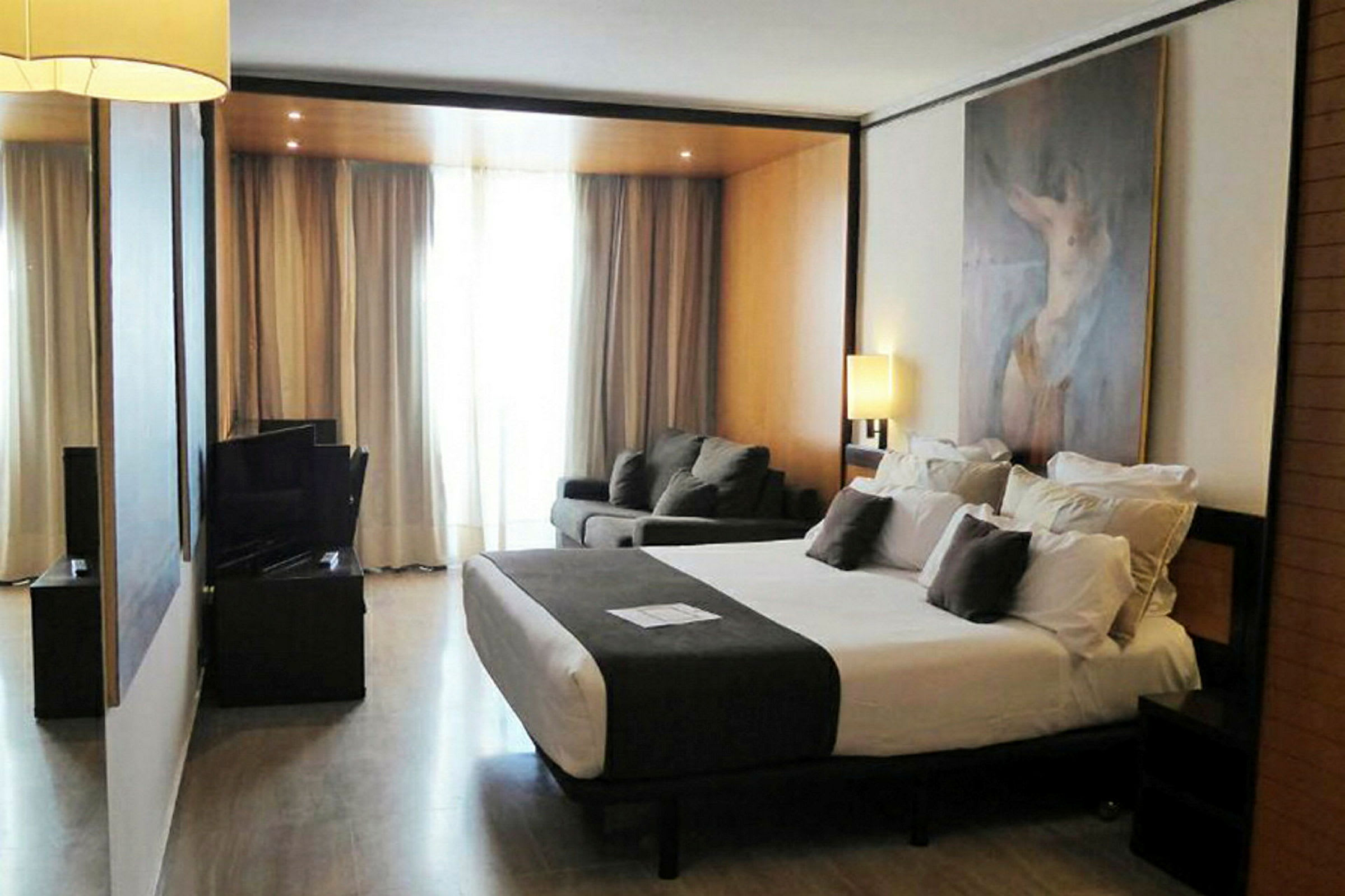 Et hotellrom med stor seng, sittegruppe og lamper, samt et maleri på veggen. (AI-generert tekst)
