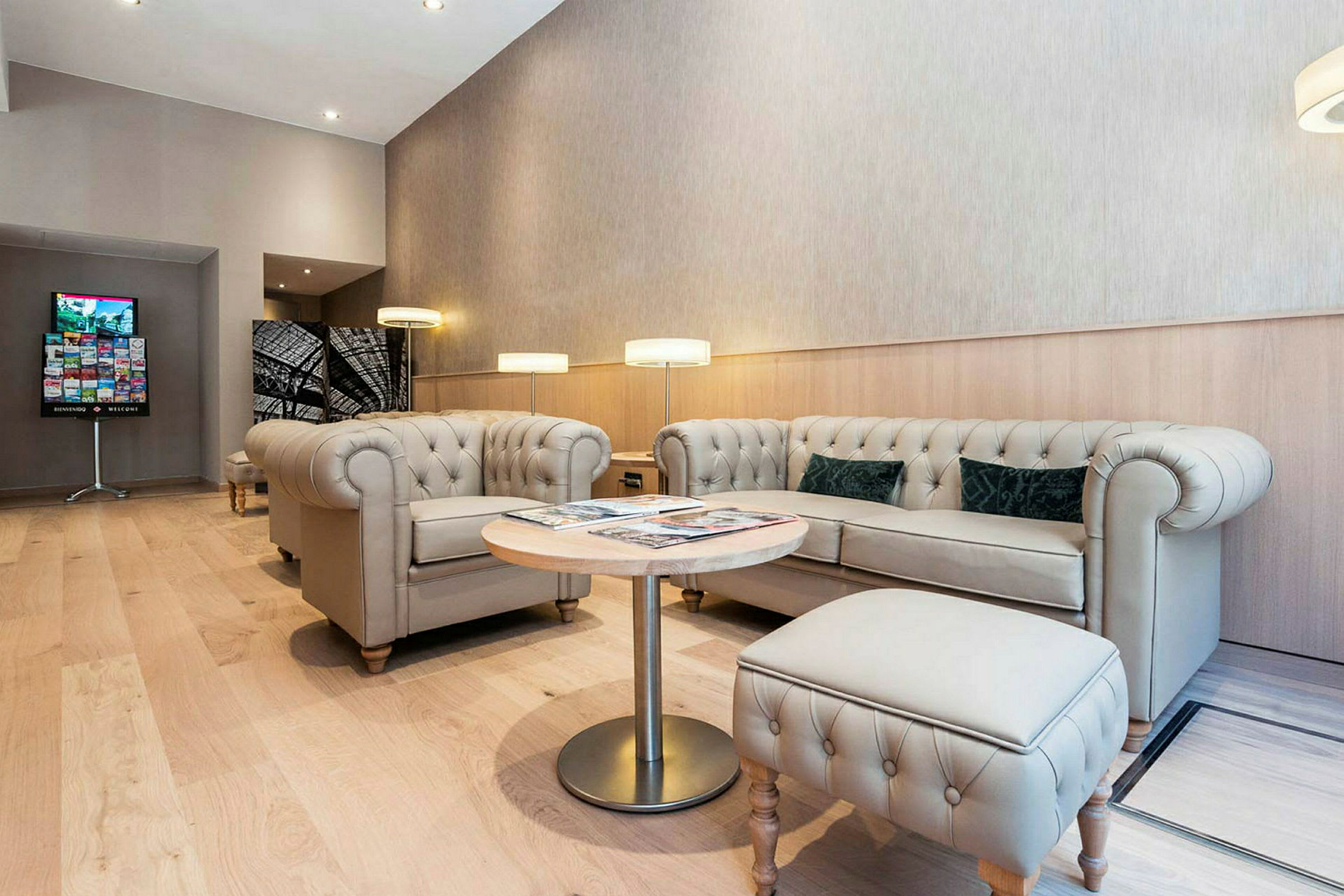 Et moderne loungeområde med beige chesterfieldsofaer og lenestoler, et rundt trebord og gulvlamper. Gulvet er av lyst tregulv. (AI-generert tekst)