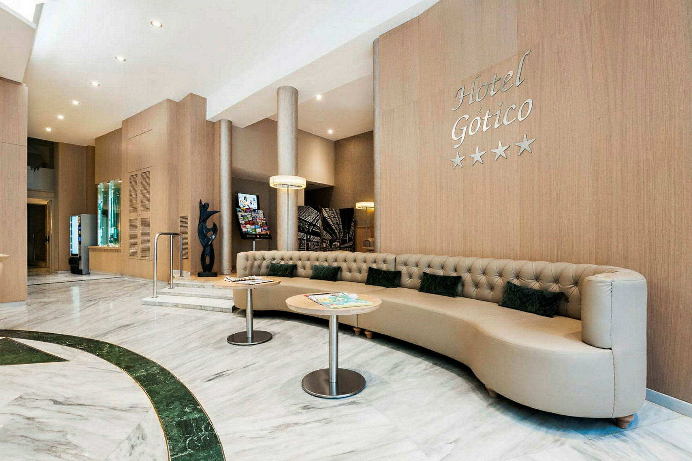 Hotellobby med moderne, lyst interiør. En stor, beige sofa med små puter og to runde bord er synlige. Veggene er dekket av trepanel, og et skilt med teksten "Hotel Gotico" ses. (AI-generert tekst)