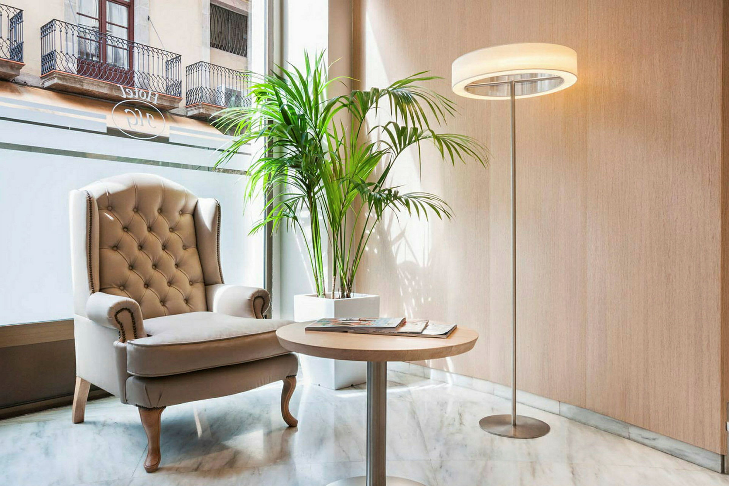 Et rom med en komfortabel beige lenestol, et rundt trebord, en lampe og en grønn plante. Store vinduer i bakgrunnen. (AI-generert tekst)