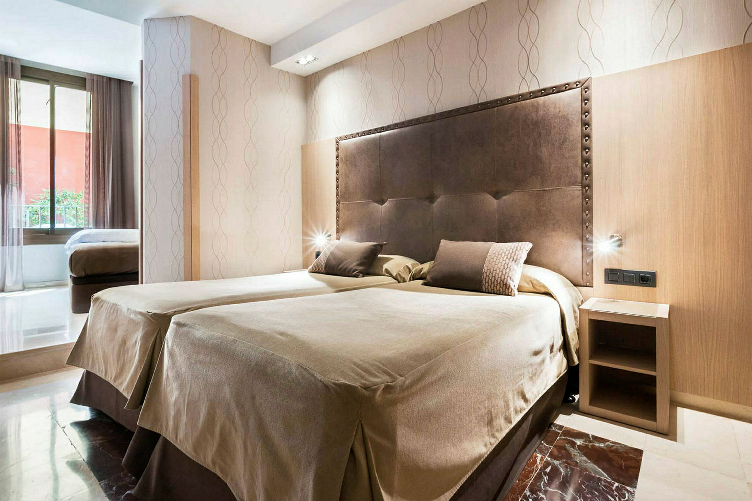 Et hotellrom med to enkeltsenger ved siden av hverandre og en sengegavl i mørkt materiale. Det er et lite nattbord og en stor vindusflate som slipper inn naturlig lys. (AI-generert tekst)