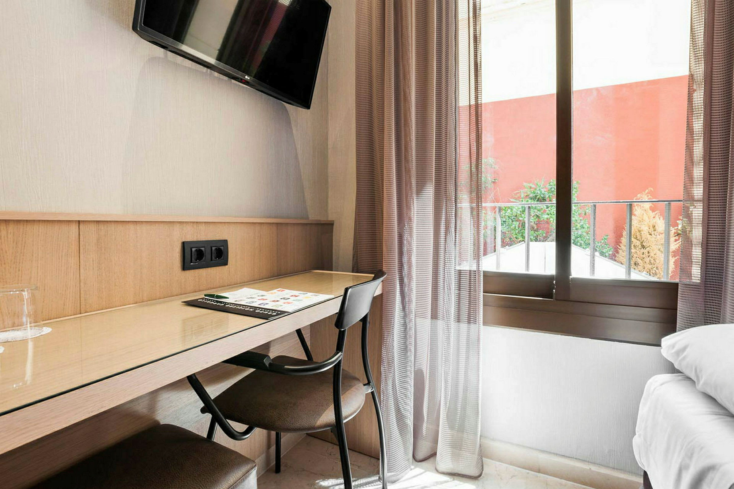 Skrivebord på et hotellrom med stol, skrivesaker, TV på veggen og et vindu i bakgrunnen. (AI-generert tekst)