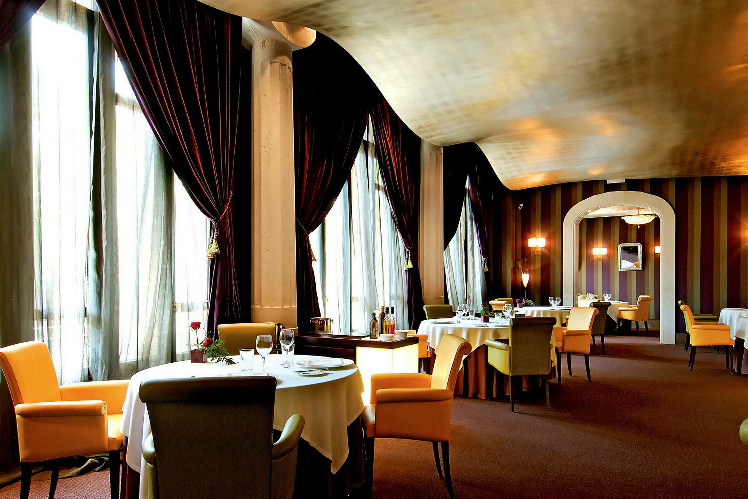 En elegant restaurant med dekkede bord og komfortable stoler, omgitt av store gardiner og dempet belysning. (AI-generert tekst)