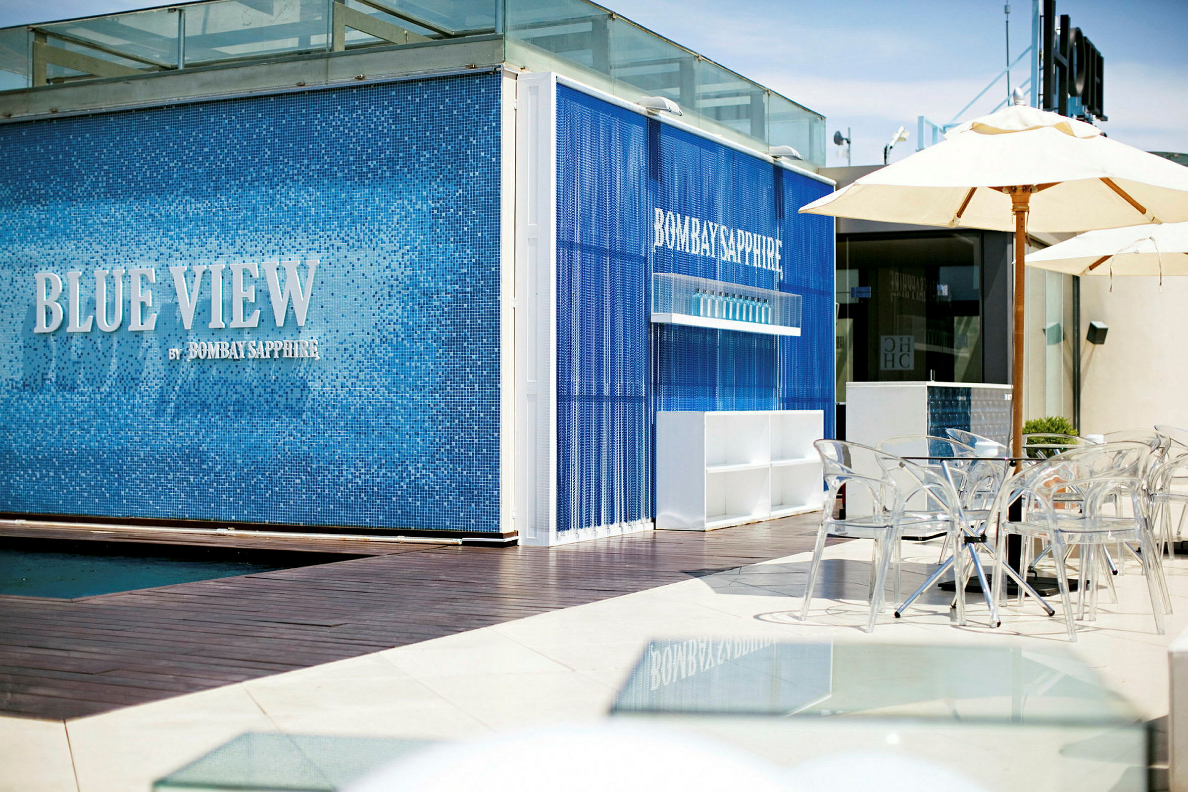 En moderne bar utvendig med blå fliser og teksten "Blue View by Bombay Sapphire". Foran er gjennomskinnelige stoler og parasoller. (AI-generert tekst)