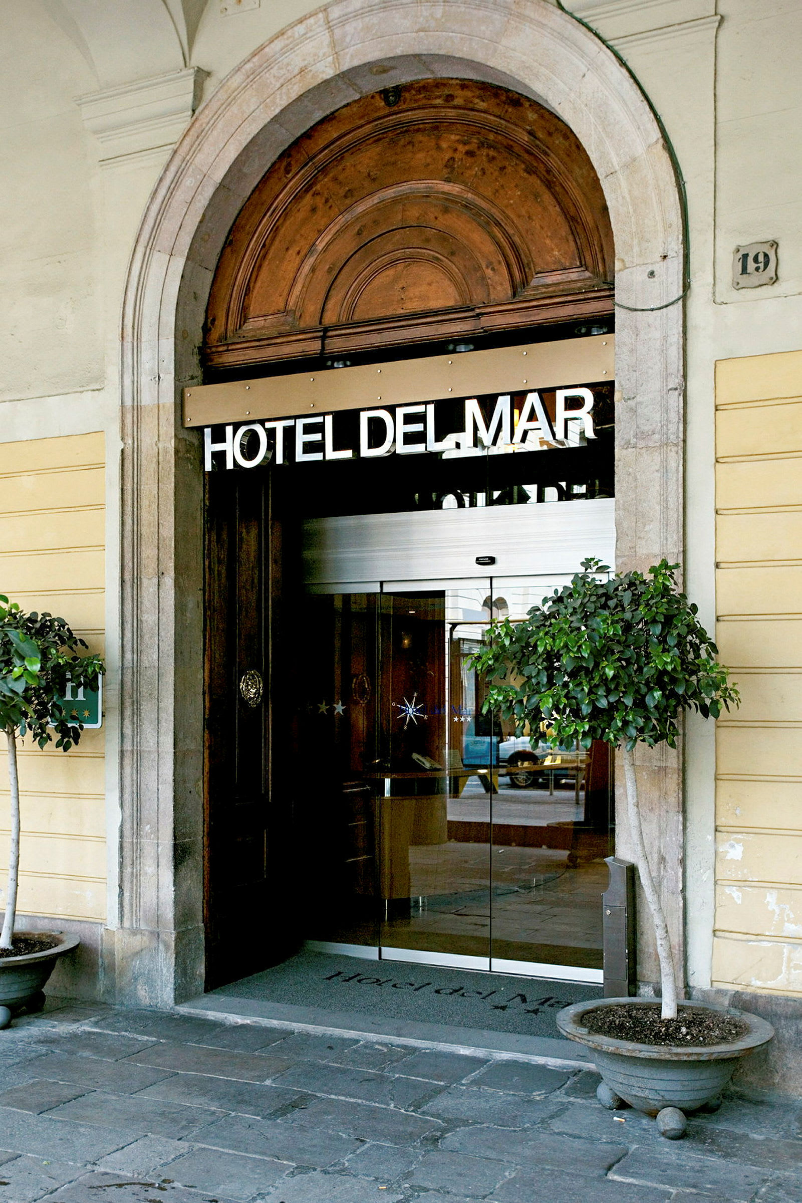 En inngang til et hotell merket med teksten 'HOTEL DEL MAR'. Inngangen er buet med glassdører og har dekorative blomsterpotter på hver side. (AI-generert tekst)