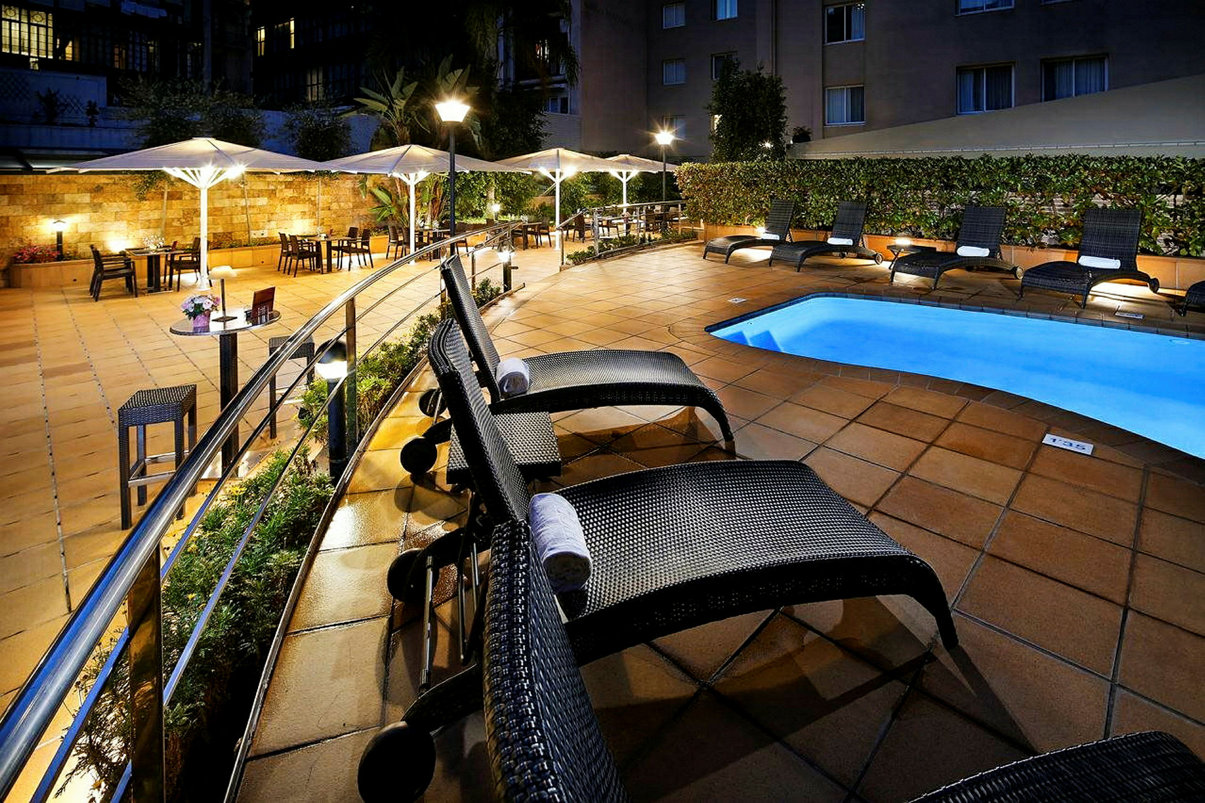 Hotellens bassengområde med solsenger og parasoller ved et opplyst flisebelagt uteområde. (AI-generert tekst)