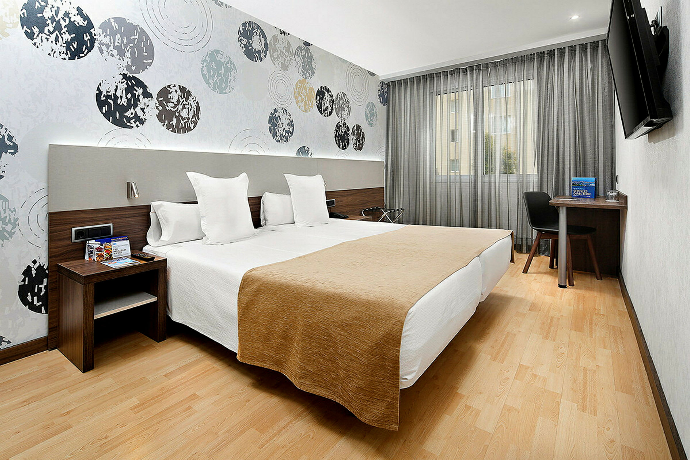 Et hotellrom med to enkeltsenger plassert ved siden av hverandre. Veggene er dekorert med et mønster av sirkler, og det finnes en TV, skrivebord og stol ved vinduet. (AI-generert tekst)