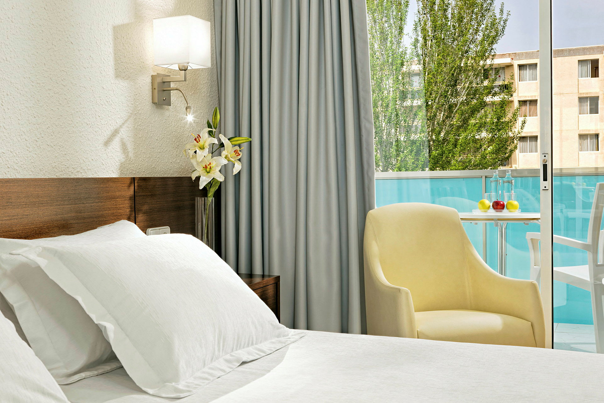 Et hotellrom med en hvit oppredd seng, en gul lenestol og et bord ved en balkong med glassrekkverk. En blomsterkrukke med hvite liljer står på et bord. (AI-generert tekst)