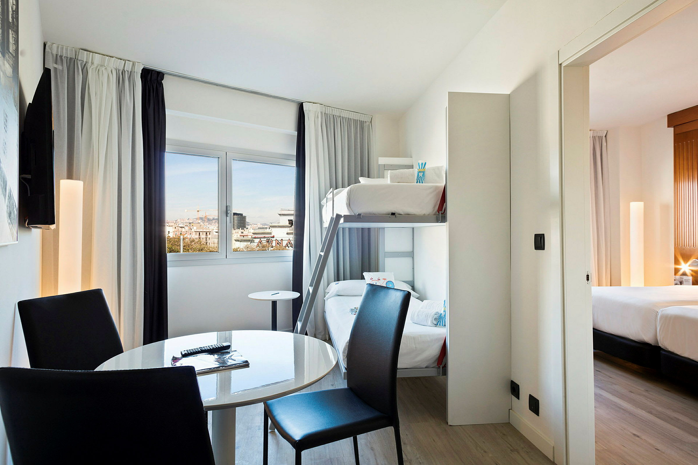Et hotellrom med køyesenger, et rundt bord og stoler, samt en utsikt gjennom vinduet. Et tilknyttet soverom med dobbeltseng vises. (AI-generert tekst)