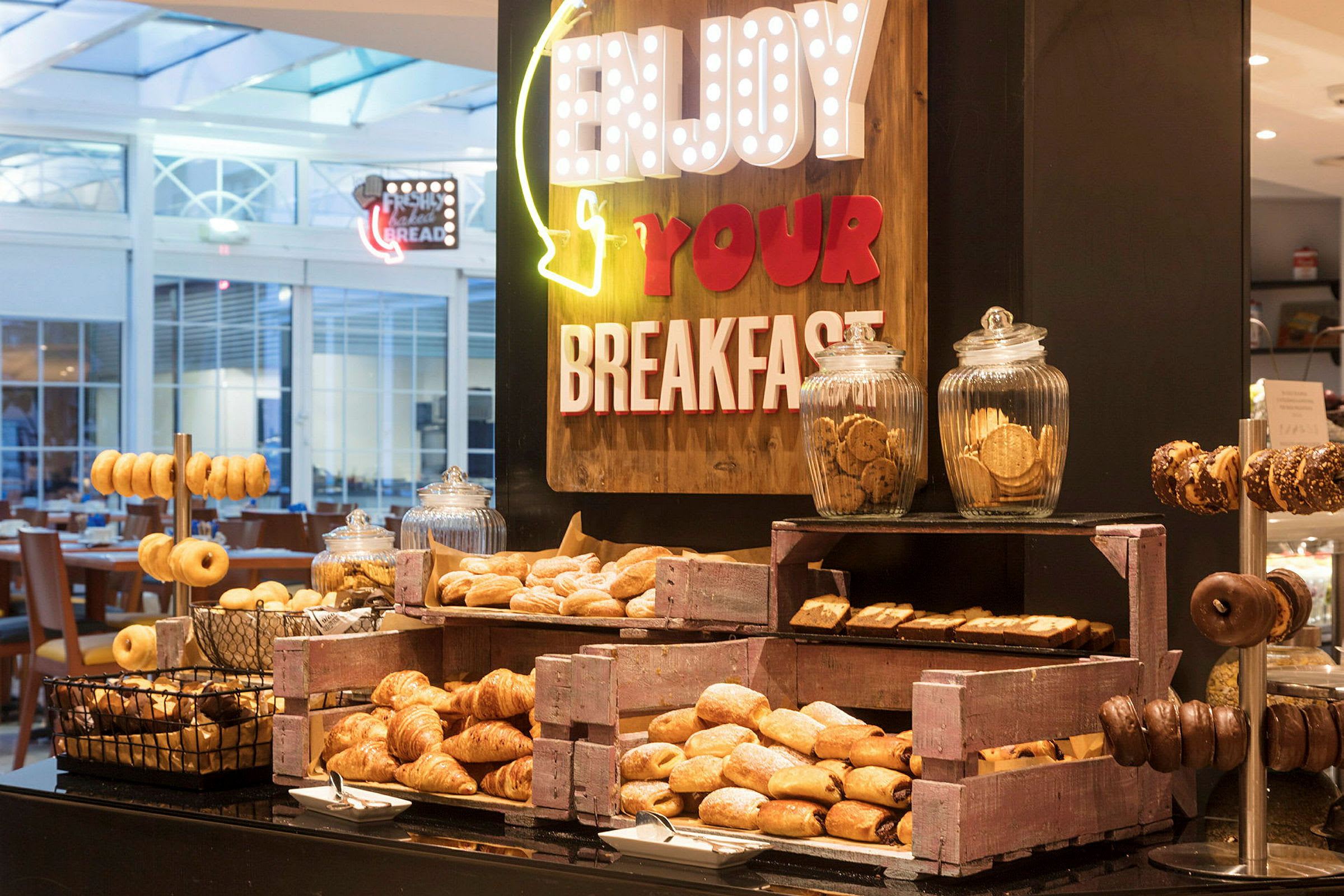 En buffé med forskjellige frokostbakverk som croissanter, donuts og småkaker, arrangert med skilt som oppfordrer til å nyte frokosten. (AI-generert tekst)