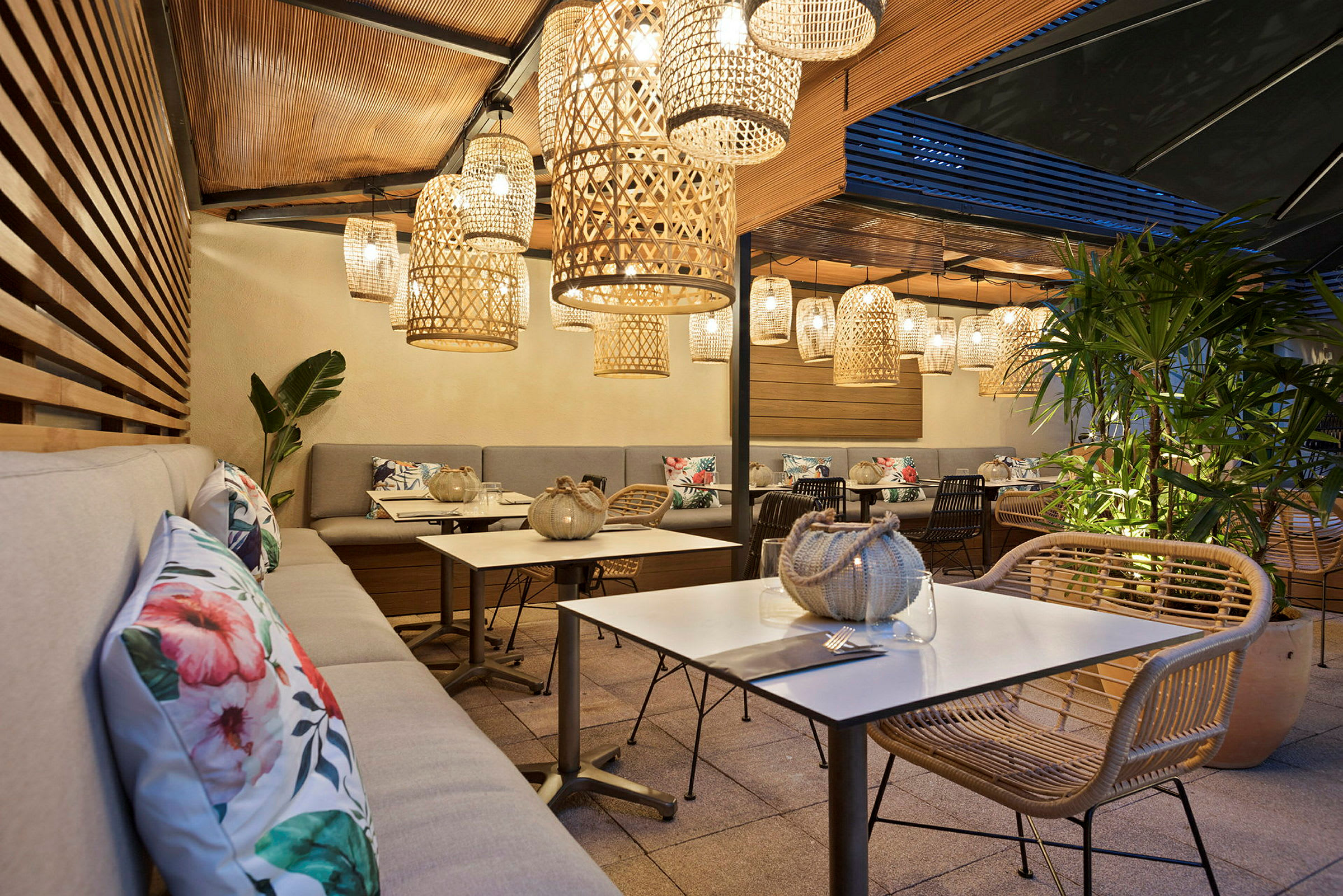 Terrasse med sitteplasser, bord og dekorative kurvlamper som henger fra taket. Planter og pynteputer er synlige. (AI-generert tekst)