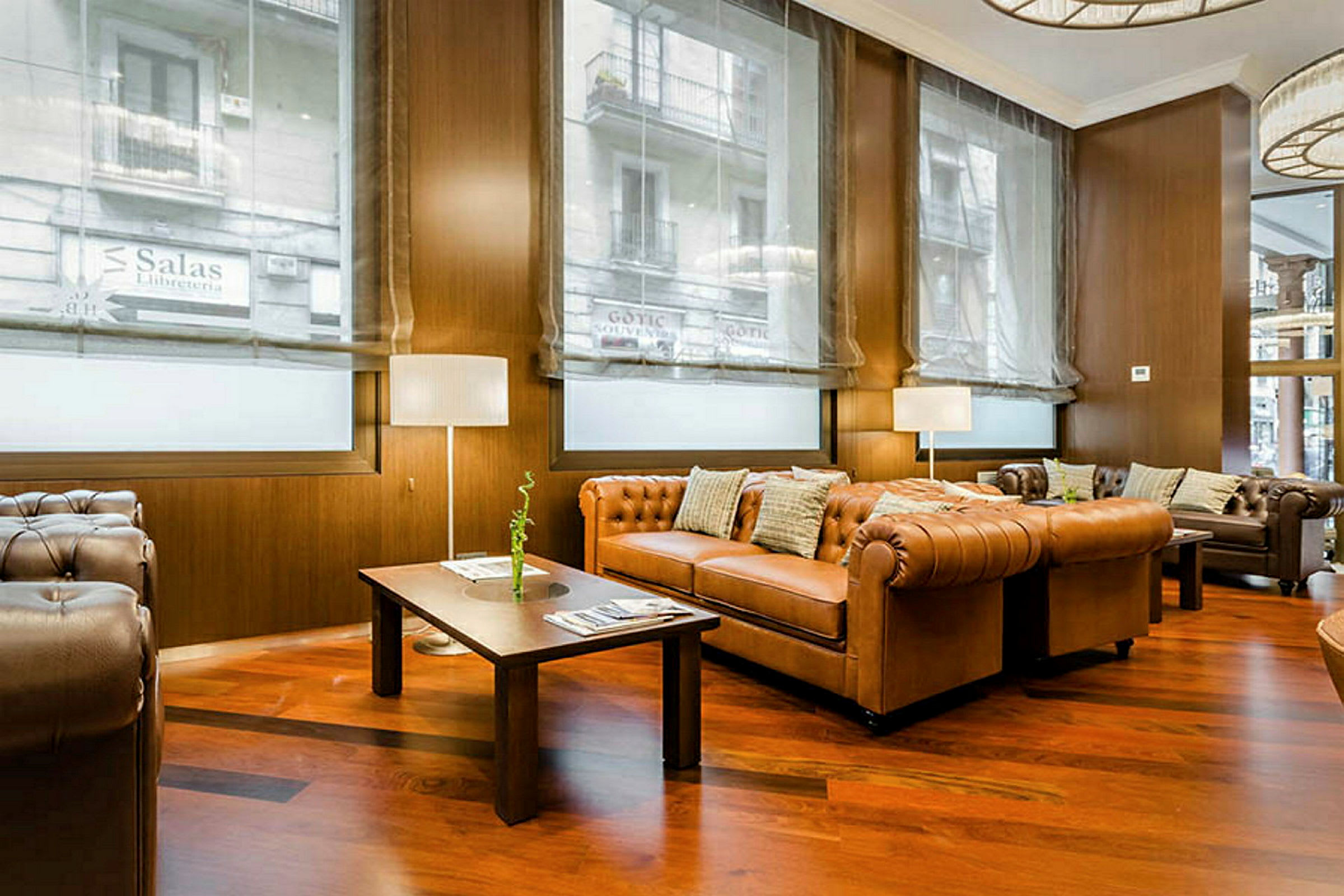 En hotellobby med brune skinnsofaer, trebord og store vinduer med gardiner som slipper inn lys. (AI-generert tekst)