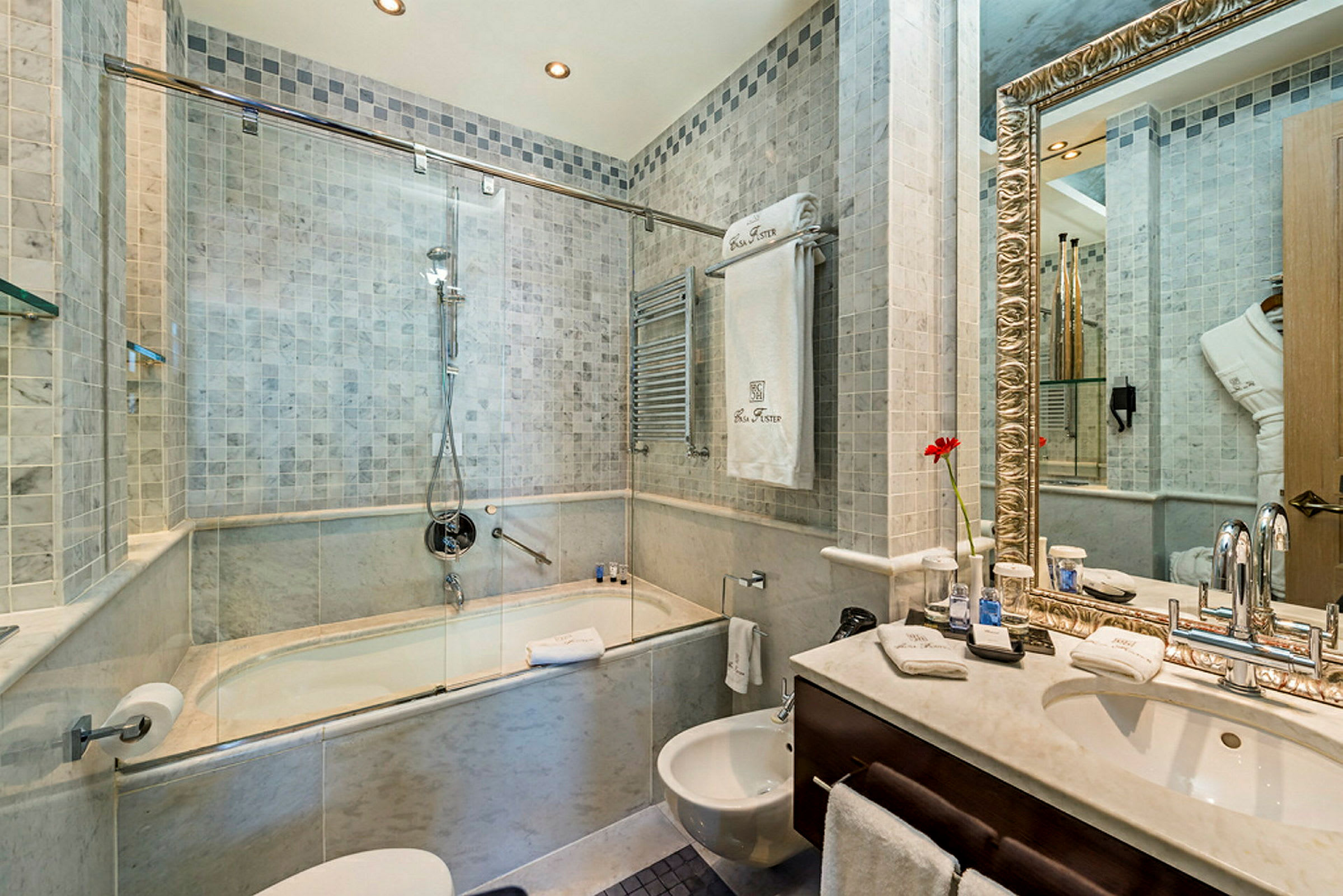 Et baderom med en dusj kombineret med badekar, en servant med et stort speil og en bidet. Veggene er flislagte, og det er håndklær og andre toalettartikler. (AI-generert tekst)