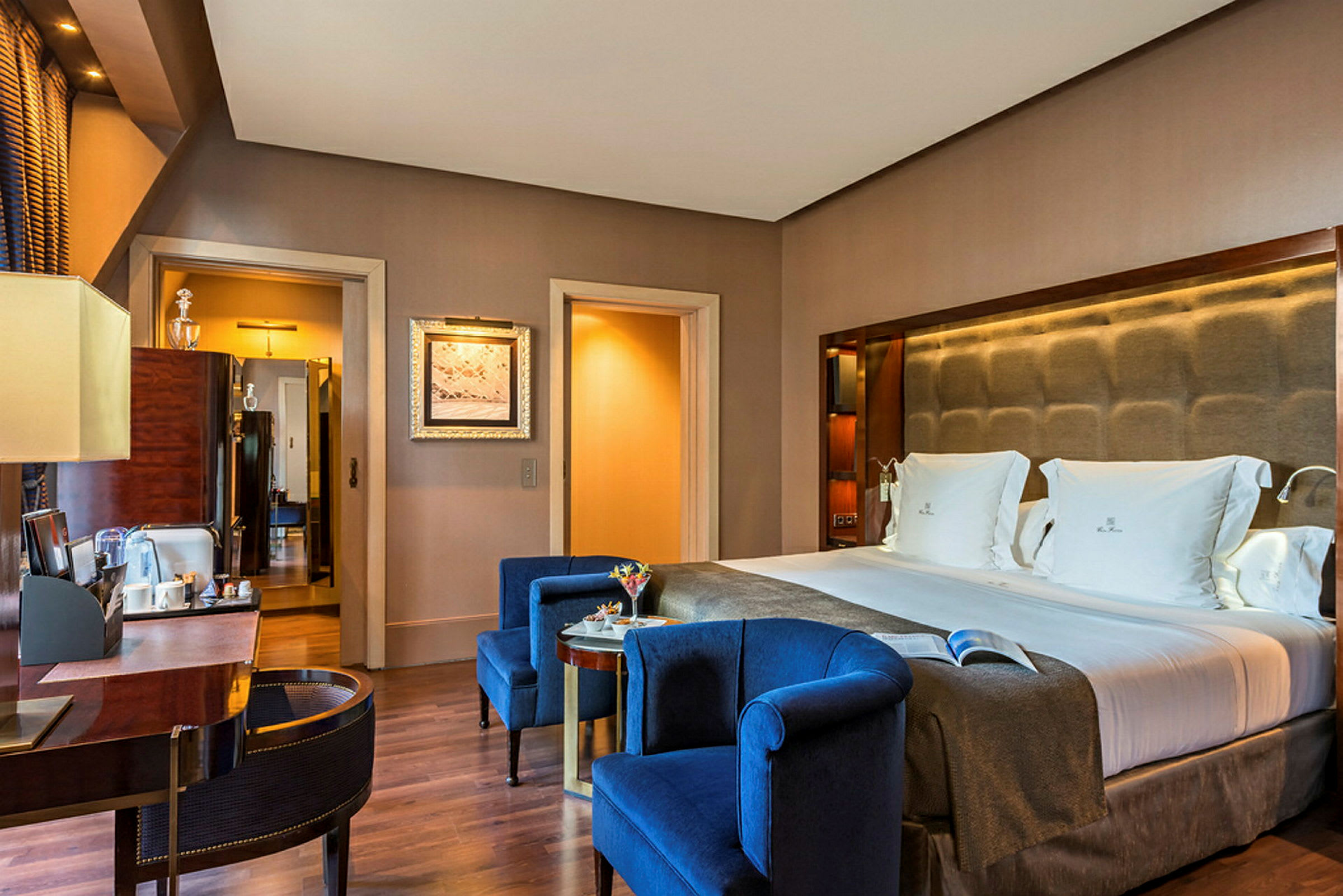 Et elegant hotellrom med en dobbeltseng og blå lenestoler foran et bord. (AI-generert tekst)