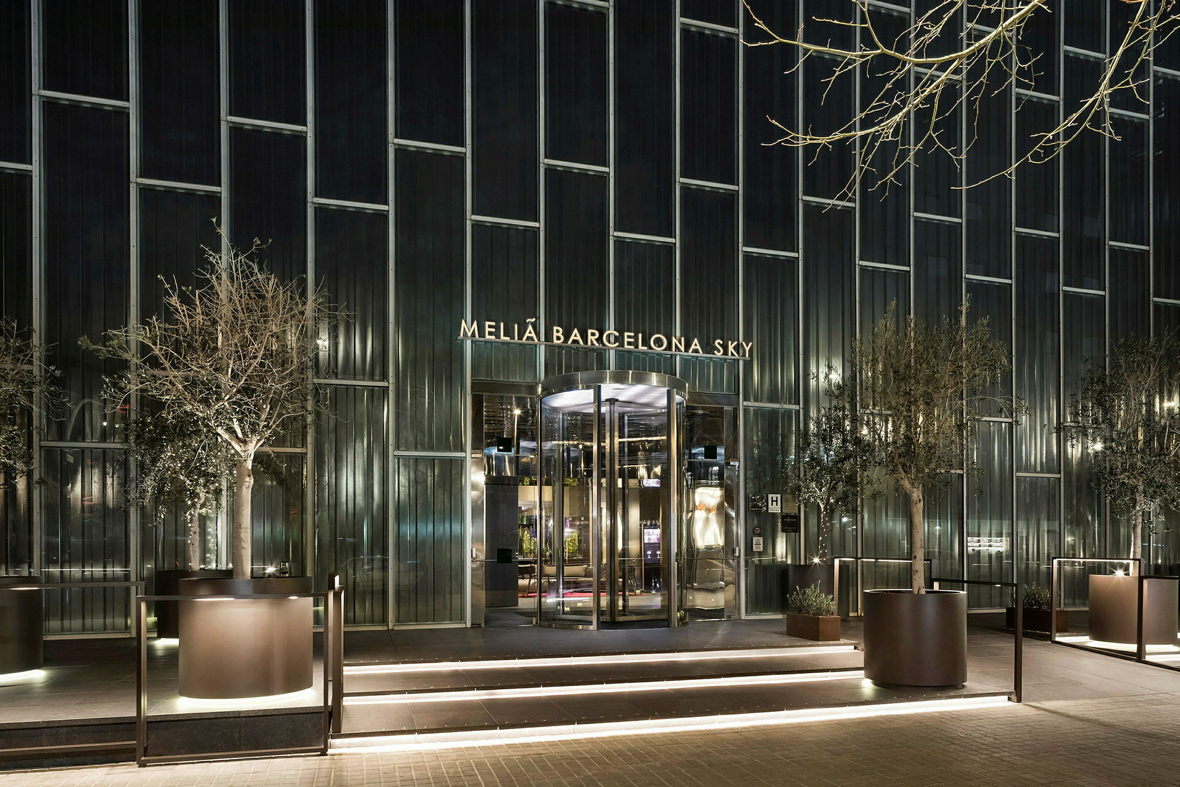 Fasaden til Melia Barcelona Sky Hotel med opplyst inngang og store glassvinduer, omgitt av planterte trær. (AI-generert tekst)