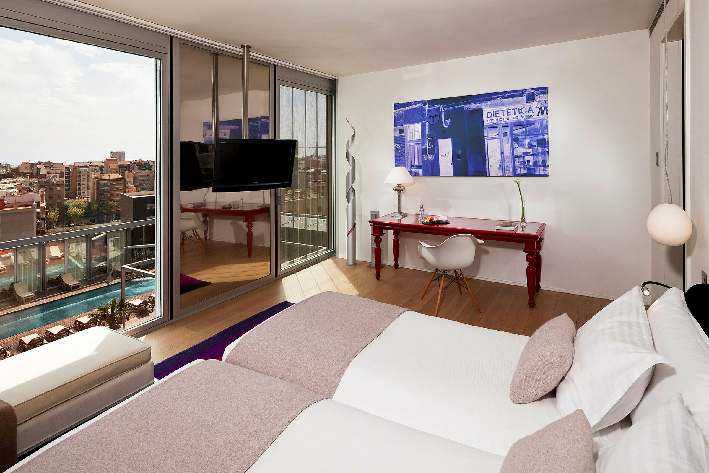 Et moderne hotellrom med to enkeltsenger, et rødt skrivebord med stol, og et stort vindu som vender mot bassengområdet og byen. (AI-generert tekst)