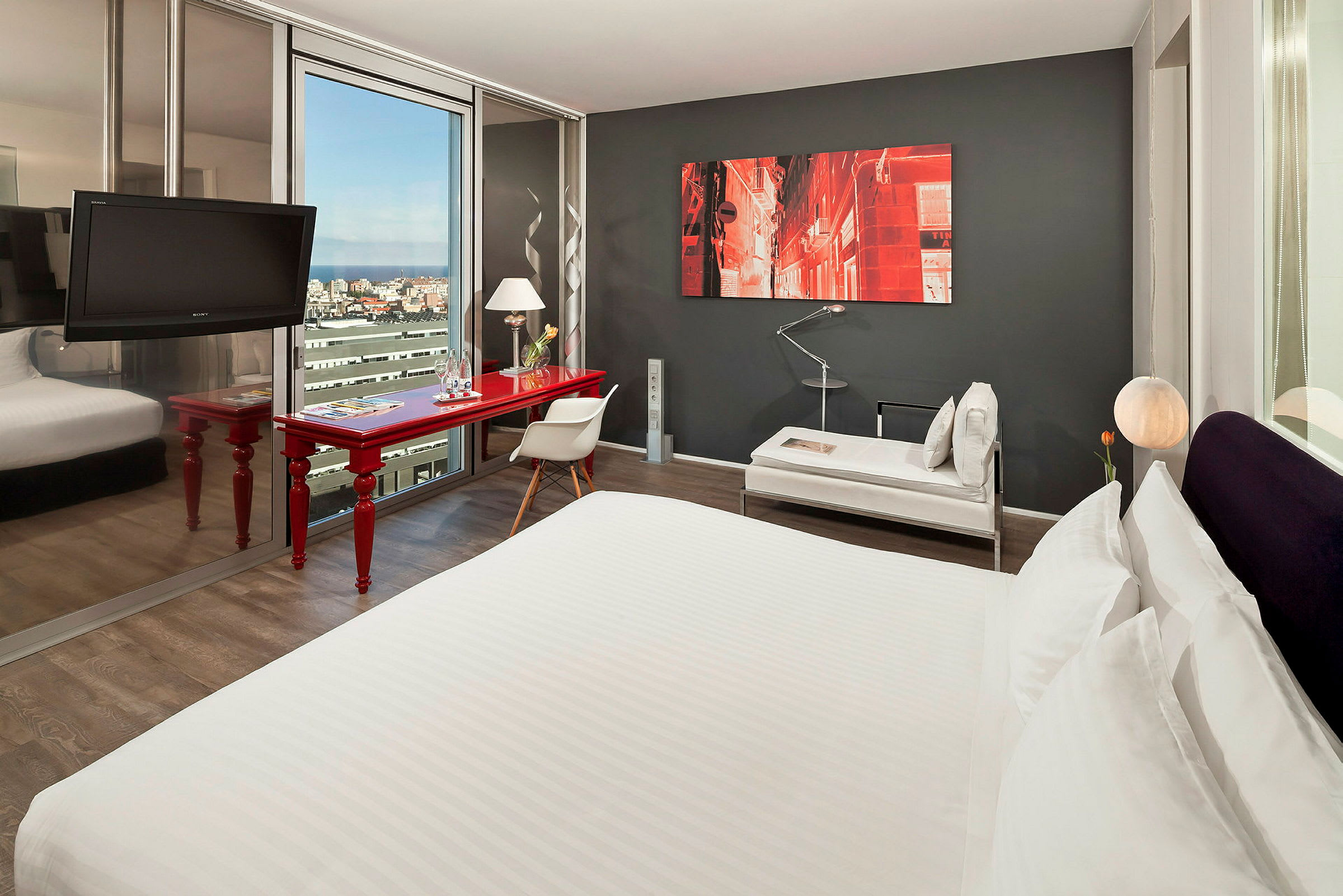 Et moderne hotellrom med en stor seng, et rødt skrivebord, en stol og et sofabord. Et maleri pryder den mørkegrå veggen, og et stort vindu viser en utsikt over byen. (AI-generert tekst)