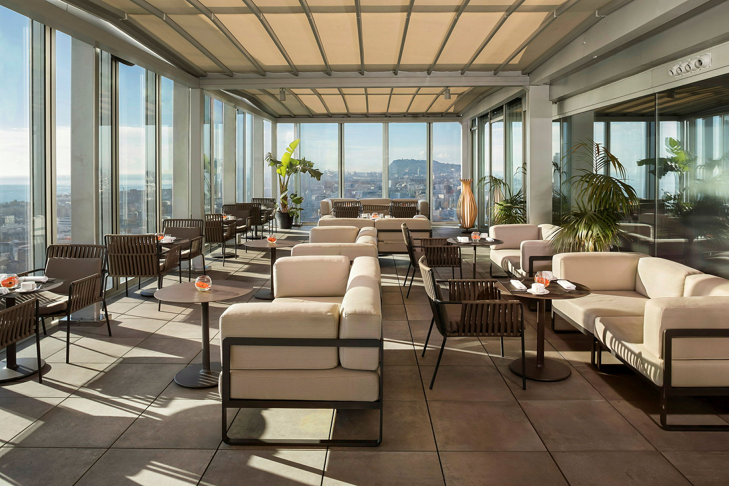 En moderne takterrasse med elegante sittemøbler og glassvegger med utsikt over en by. Solrikt vær og grøntområde gir en behagelig atmosfære. (AI-generert tekst)