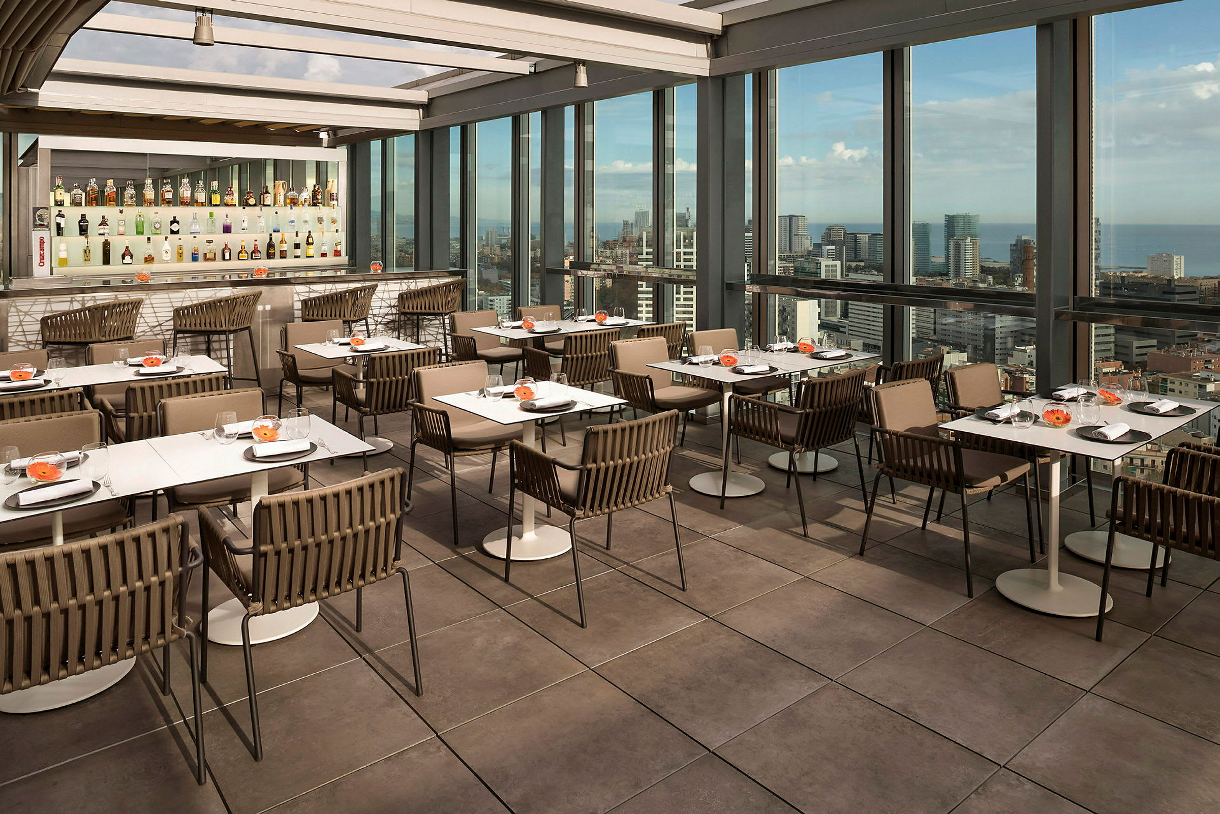 En restaurant med bord og stoler på en terrasse med store glassvinduer som viser en byutsikt. (AI-generert tekst)