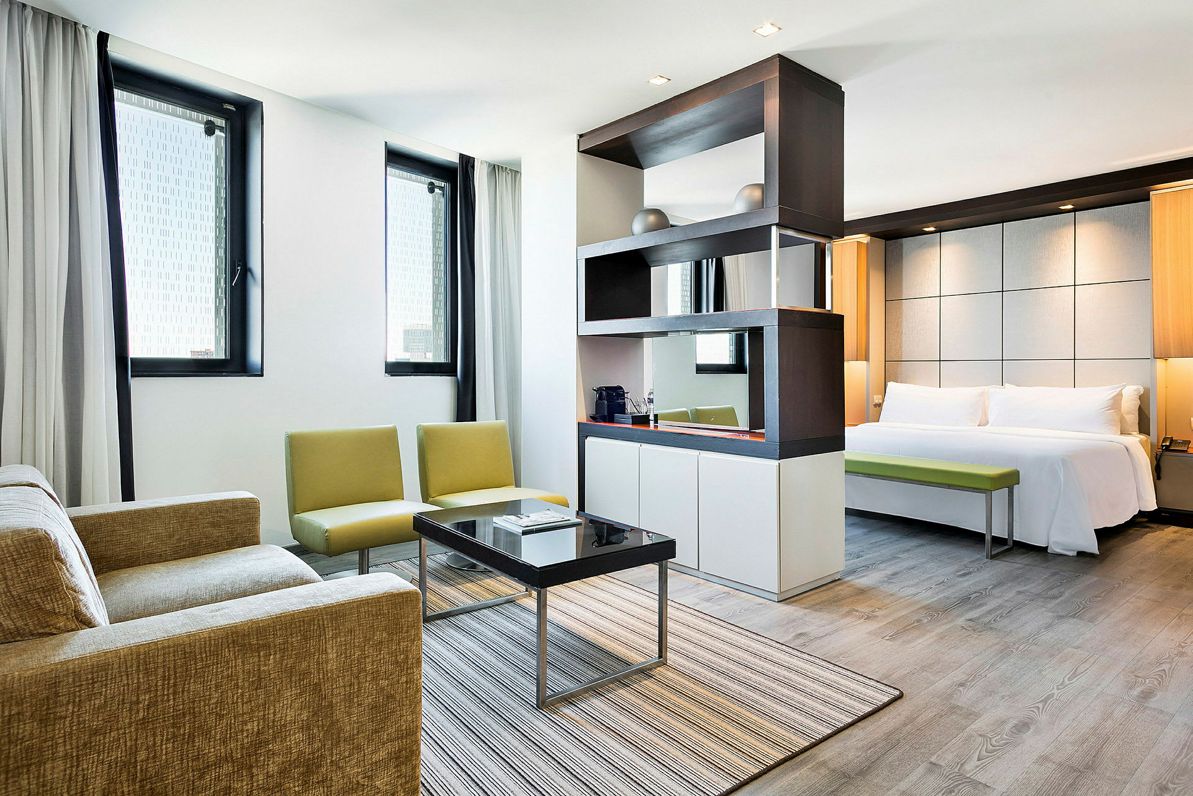 Et moderne hotellrom med en stue og et soverom. Stuen har sofa, lenestoler og sofabord. Soverommet har en dobbeltseng og en benk. (AI-generert tekst)