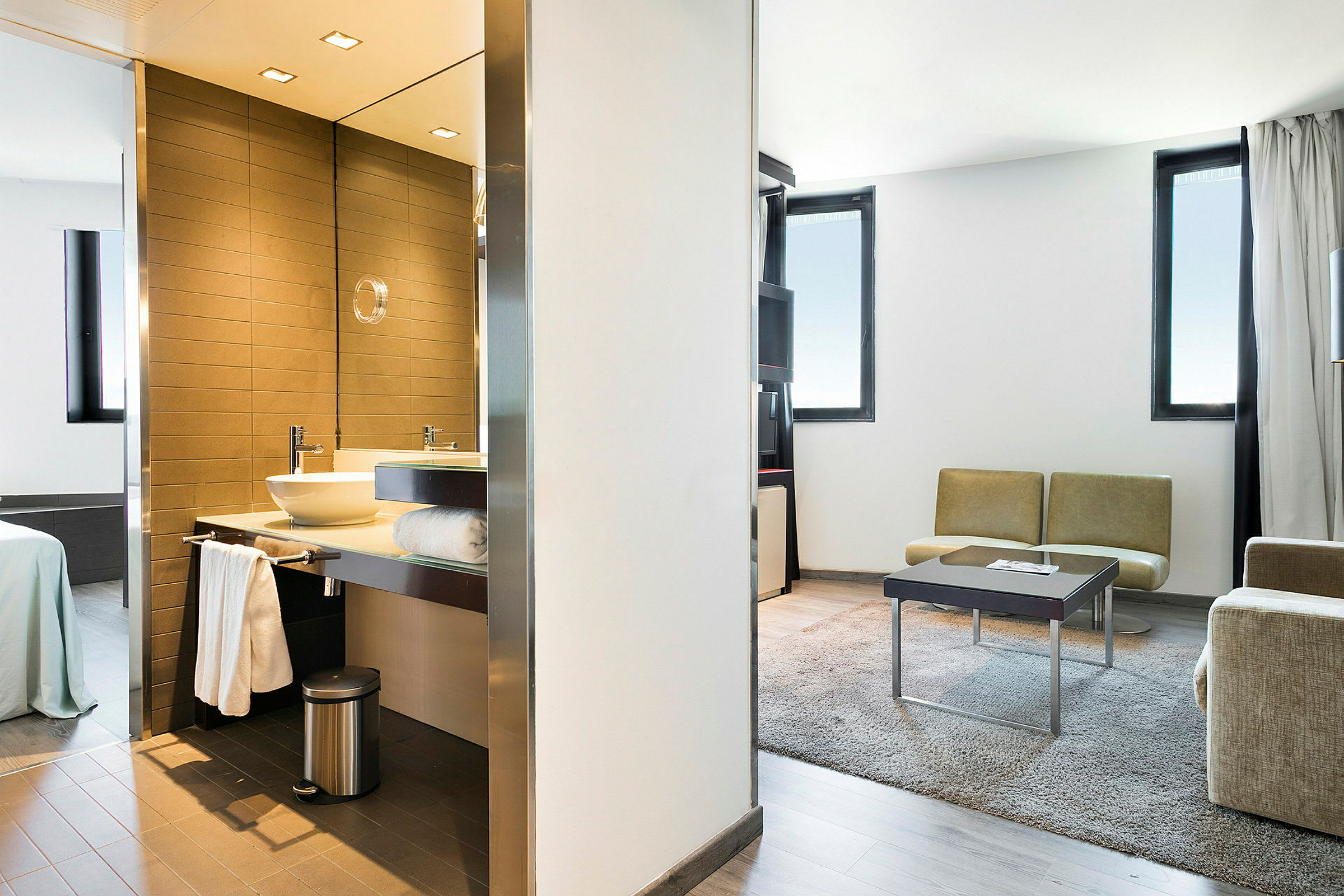 Et moderne hotellrom som viser et bad med servant og speil til venstre, soverommet med seng lenger bak, samt stuen med sofaer, bord og teppe til høyre. (AI-generert tekst)