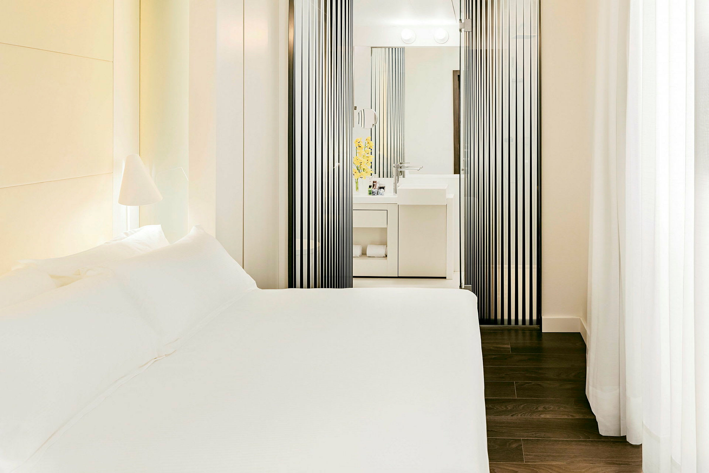 Et hotellrom med en hvit seng og et moderne bad synlig gjennom vertikale svarte paneler. (AI-generert tekst)