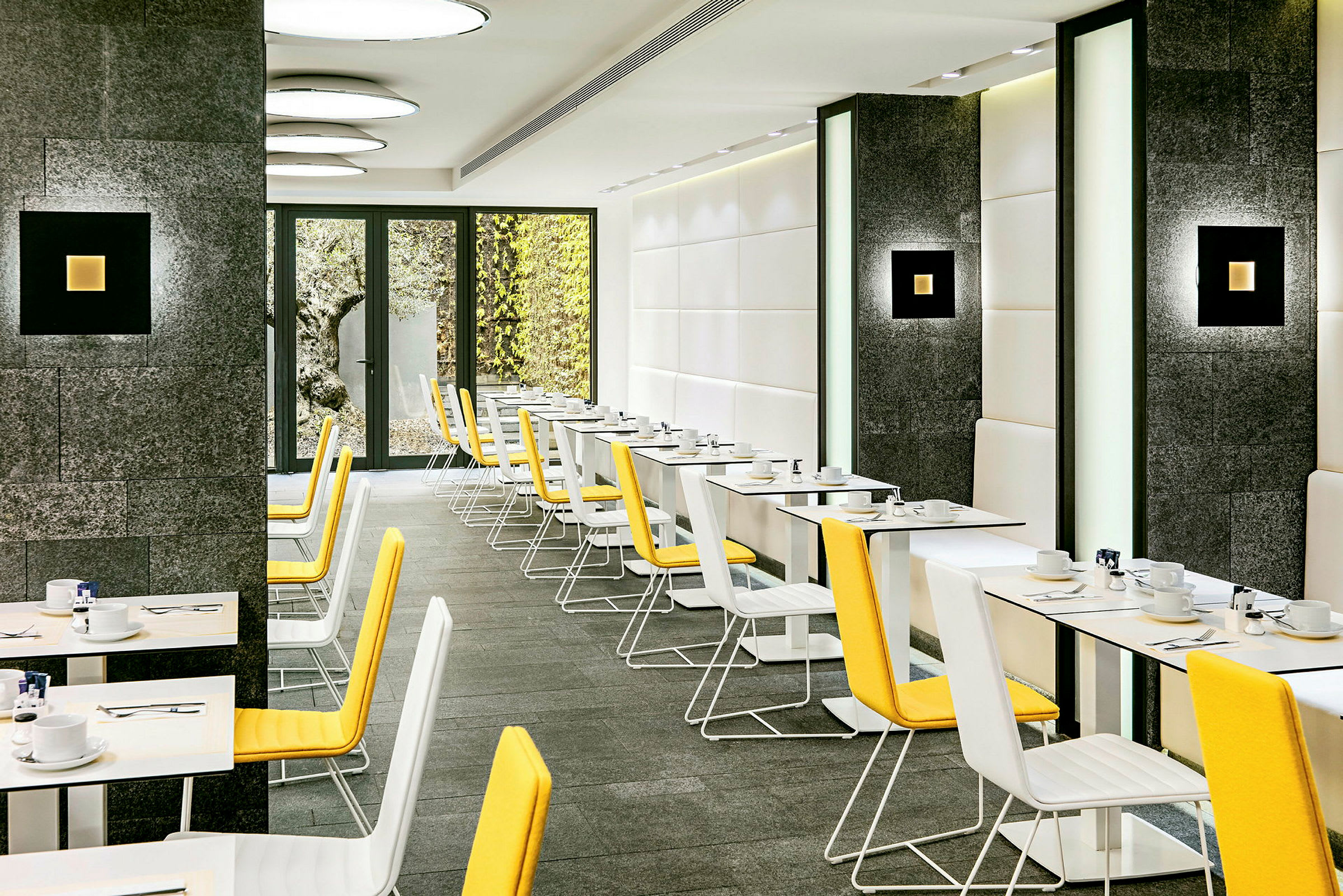 En moderne restaurant med hvite vegger og mørke steinpaneler. Gulstoltrukne stoler omgir hvite bord med porselen, bestikk og kopper plassert. Glassdører fører til en bakgård. (AI-generert tekst)