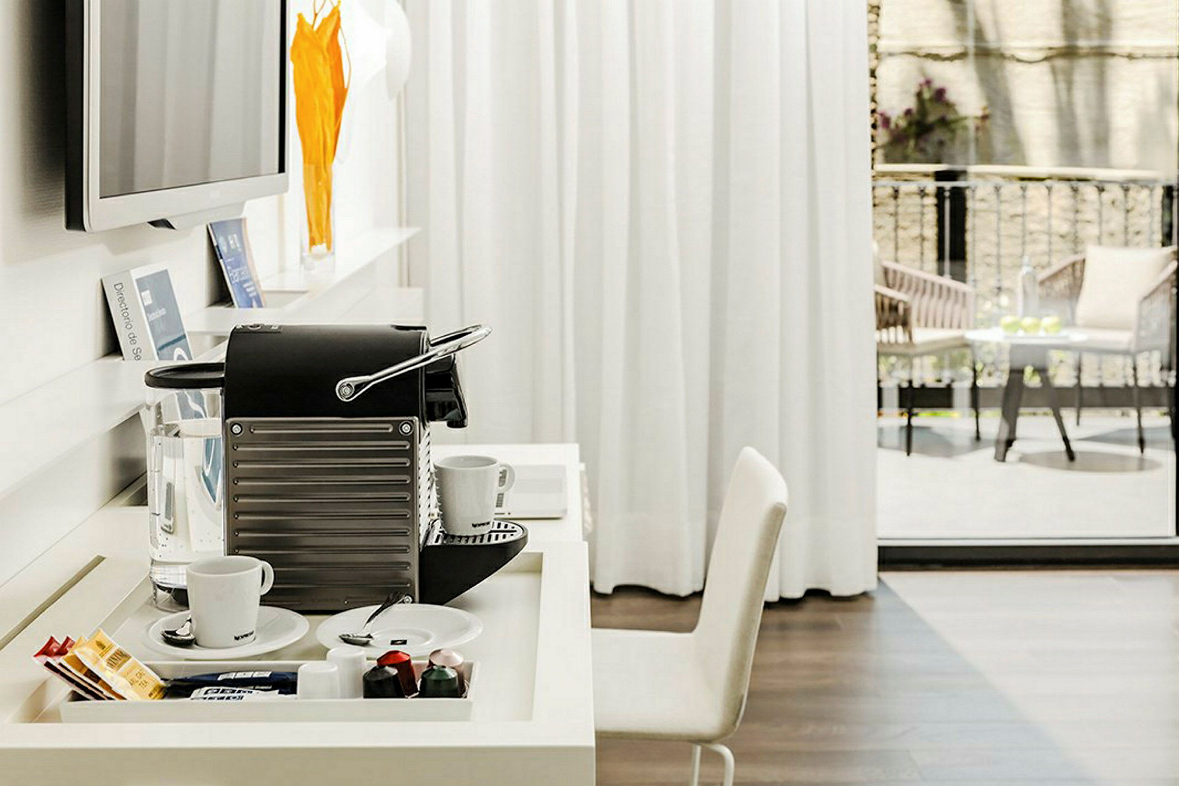 Et bord med en kapsel kaffemaskin, to kopper og kaffetilbehør står i et lyst rom. En balkong med sittemøbler er synlig gjennom glassdørene. (AI-generert tekst)