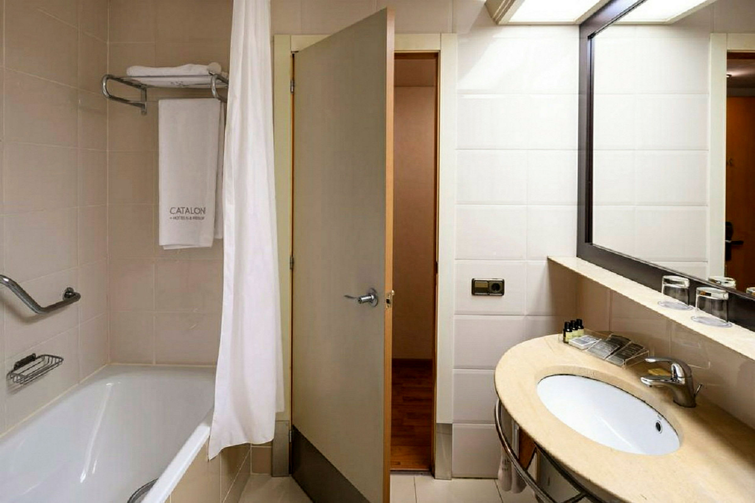 Hotellbad med et badekar med dusjforheng, et håndkle hengende på en stang og en servant med speil. (AI-generert tekst)
