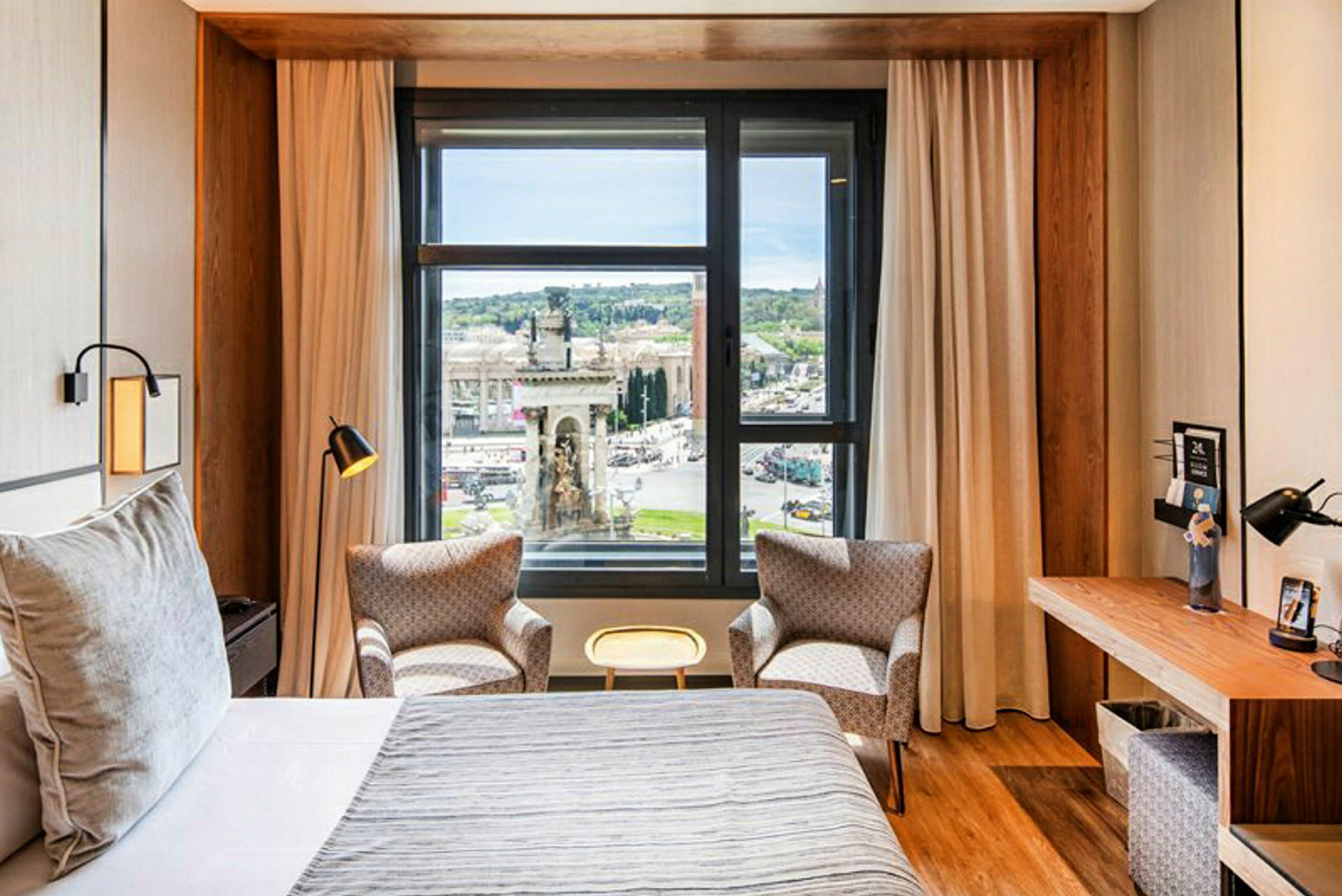 Et hotellrom med en stor seng, to lenestoler foran et vindu med utsikt over en by, samt et skrivebord på siden. (AI-generert tekst)
