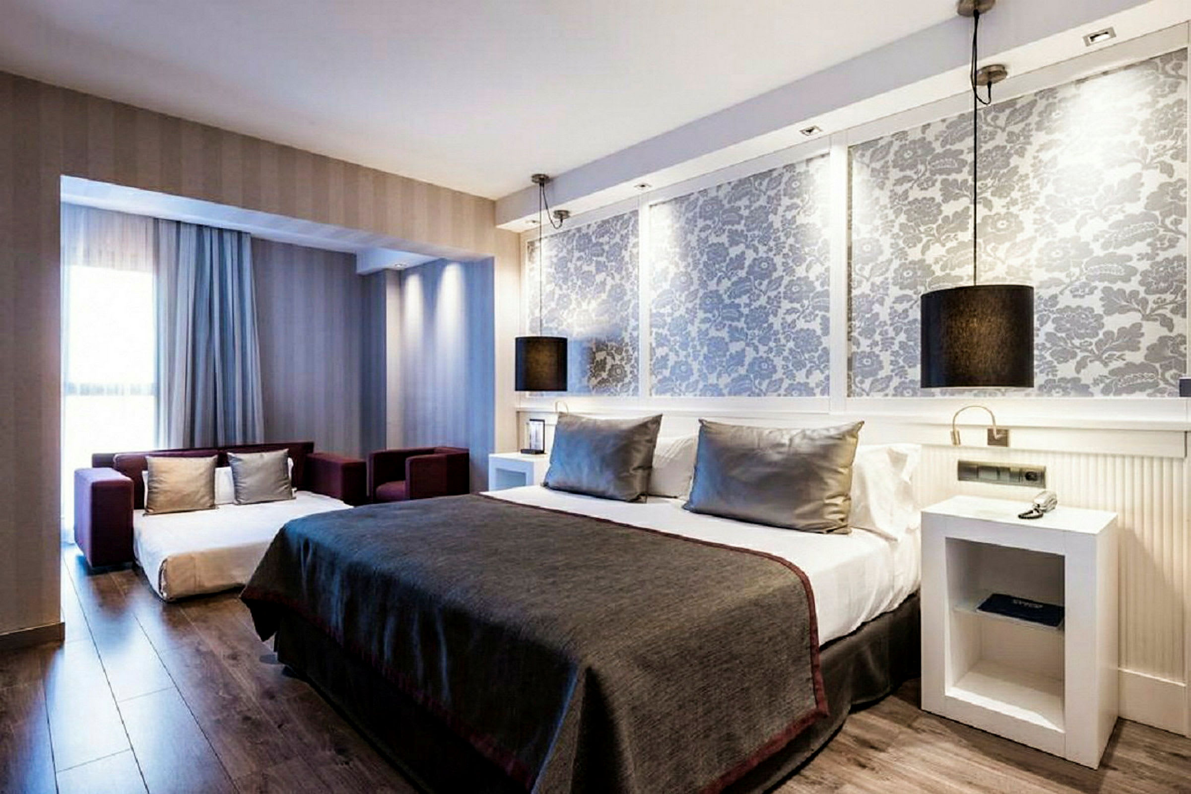 Et moderne hotellrom med grå innredning, en stor seng med grå sengeteppe og puter, samt en liten sittegruppe foran et stort vindu. (AI-generert tekst)