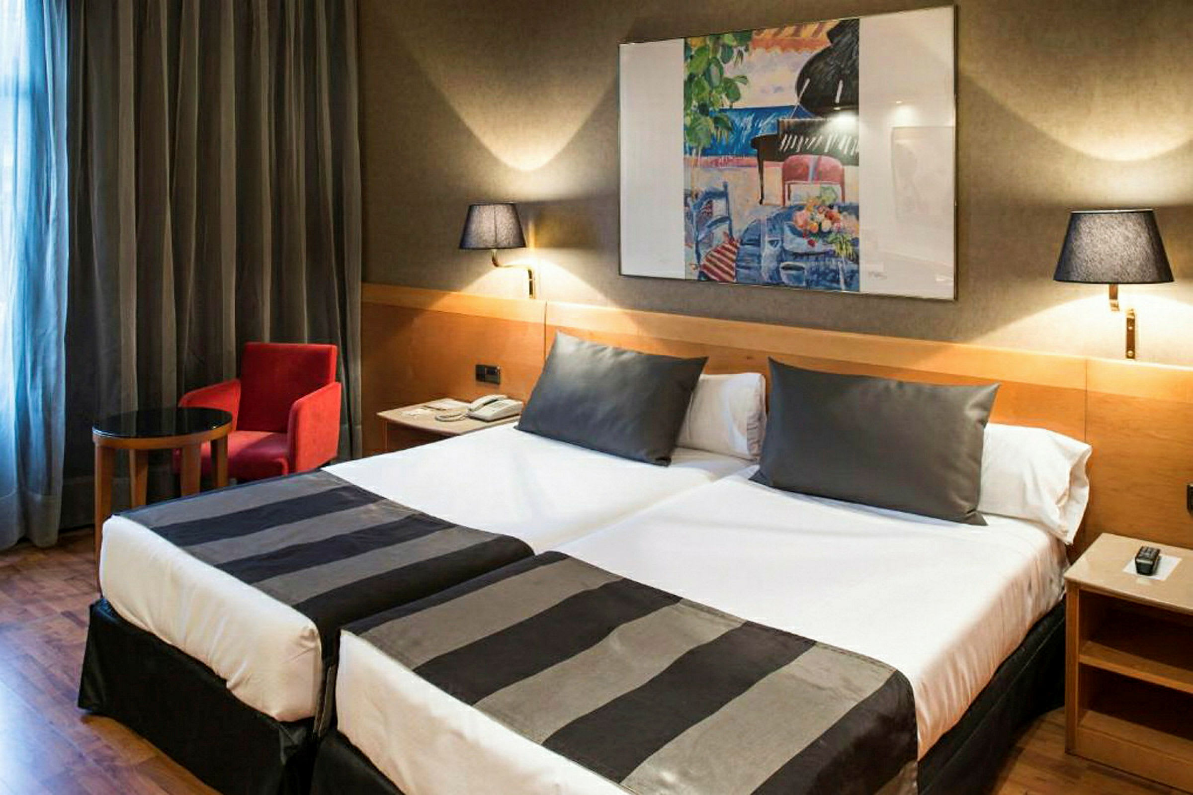 Et hotellrom med to enkeltsenger, begge oppredd med svart-hvite sengetepper. På veggen henger et fargerikt maleri, og rommet har også et lite bord, en rød lenestol og to vegglamper. (AI-generert tekst)